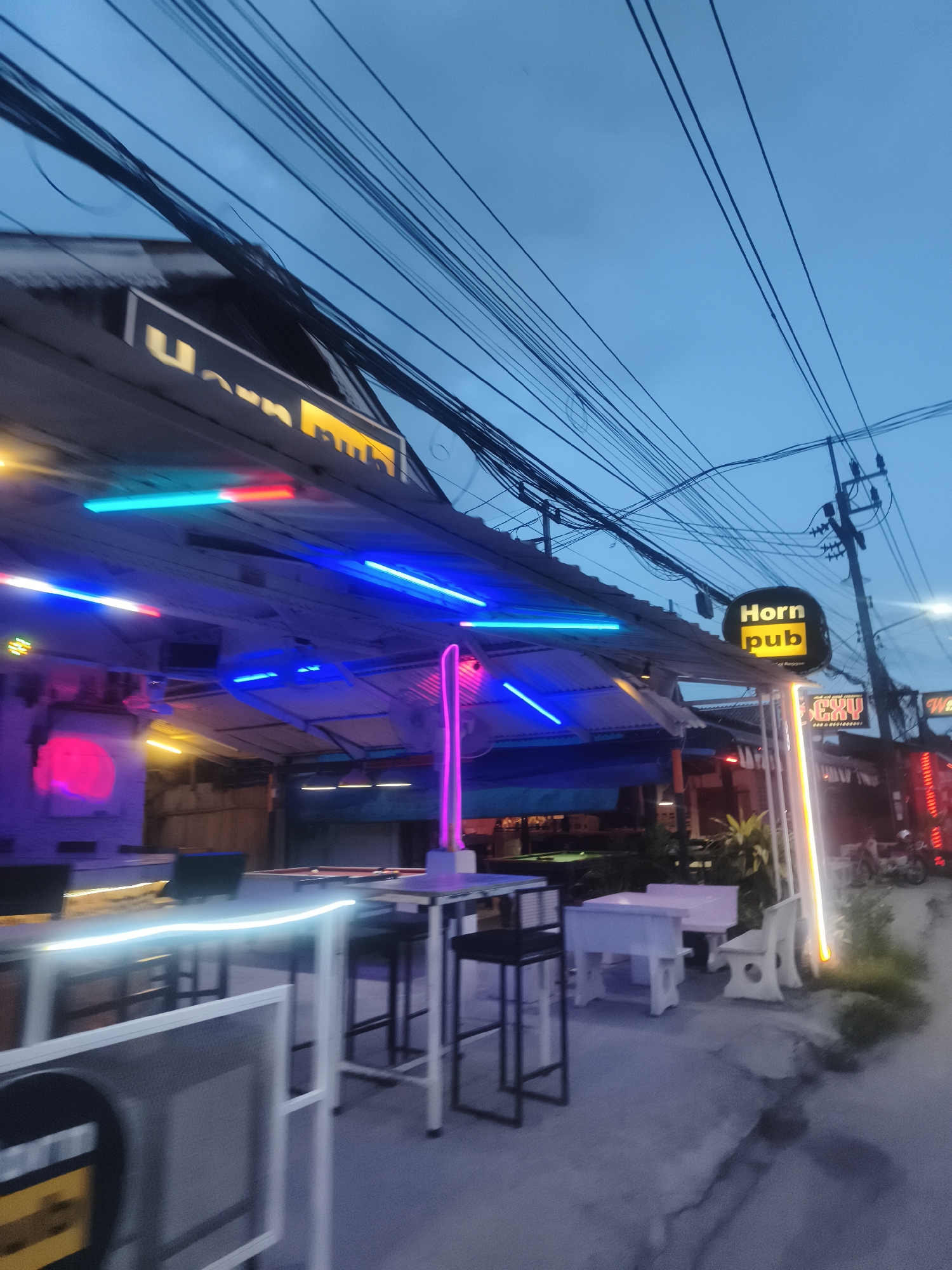 This is an exterior shot of the "Horn Pub" in Baan Chaweng, Thailand, taken at dusk. The pub is illuminated with neon lights of various colors, including blue, pink, yellow, and red. There are tables and chairs set up outside the establishment, some of which are lit with LED strips. Power lines are visible overhead against the evening sky. Other signs and storefronts can be seen further down the street. A pool table is visible inside the bar. The street and sidewalk in front of the pub are visible.