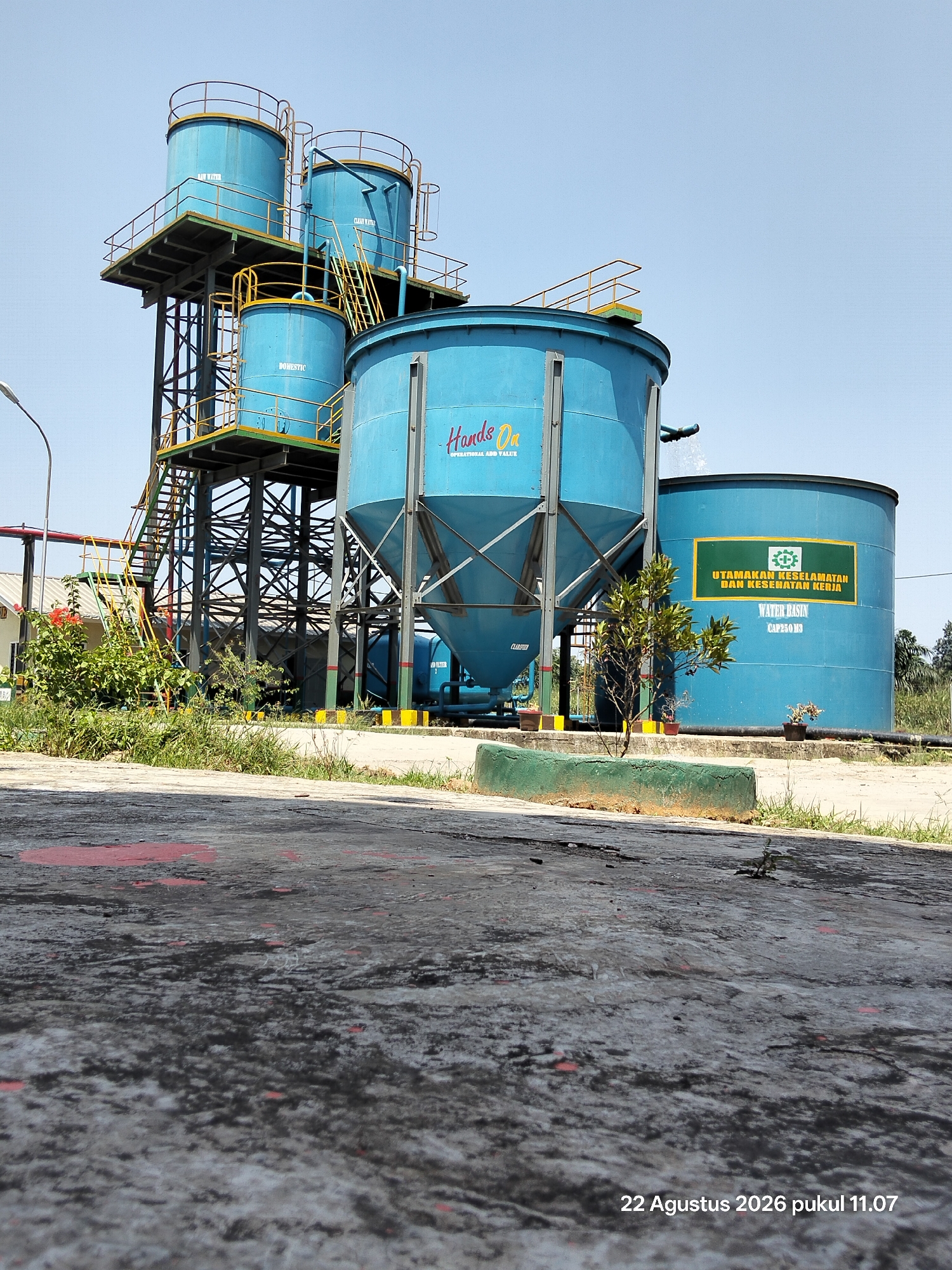 The image shows an industrial water treatment facility in Unknown, Indonesia. The foreground is dominated by a cracked and weathered concrete surface with patches of red paint. In the background, a series of large, blue cylindrical water tanks are arranged. The tanks have metal platforms and railings, with visible staircases connecting them. Some of the tanks have text labels. One large, conical tank in the center has the text "Hands On" and "OPERATIONAL ADD VALUE" in red and yellow. Another large cylindrical tank on the right has a green sign with yellow text that reads "UTAMAKAN KESELAMATAN DAN KESEHATAN KERJA" and below it, "WATER BASIN CAP 250 M3". A small, leafy tree stands in front of this tank, next to a green planter. The scene is set against a clear blue sky. The image is timestamped "22 Agustus 2026 pukul 11.07" in the bottom right corner.