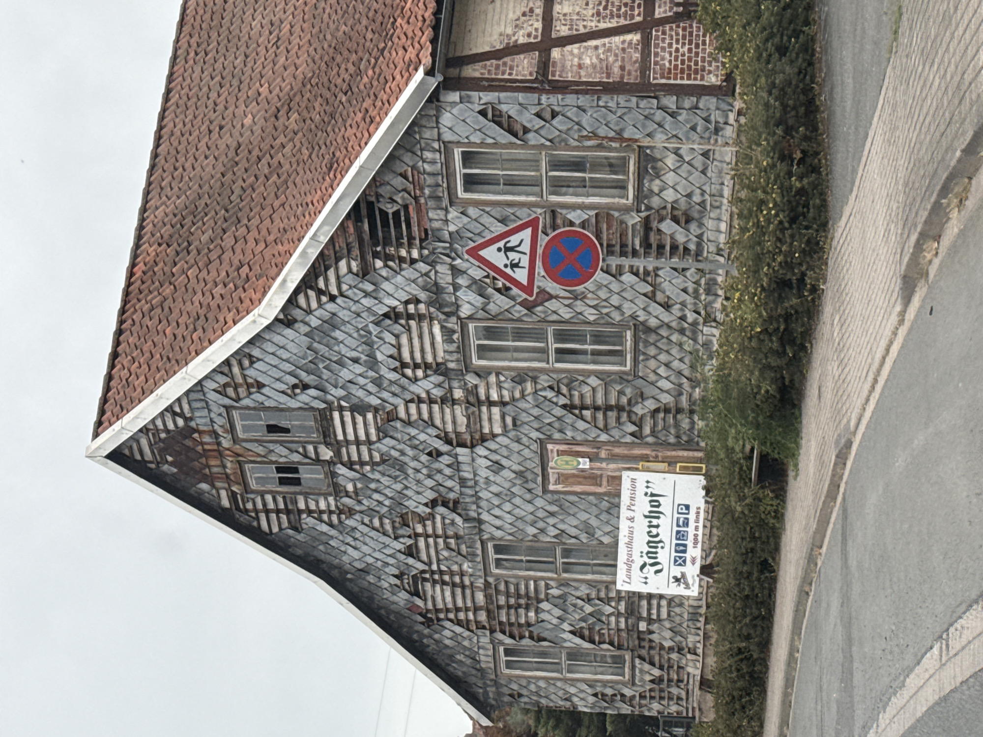 The image shows a weathered building in Ellrich, Germany, which appears to be a "Landgasthaus & Pension" named "Jägerhof". The building has a distinctive facade covered in diamond-shaped grey shingles, many of which are missing or damaged, revealing the underlying wooden structure and insulation. The roof is made of red tiles and shows signs of age.

There are several windows on the building, some of which are broken. A wooden door is visible on the left side of the building. In front of the building, there is a sign that reads "'Landgasthaus & Pension' 'Jägerhof'" with some smaller icons below it and text that appears to say "1000 m links".

Two traffic signs are mounted on a pole in front of the building. The top sign is a triangular red-bordered sign with two stick figures walking, indicating a pedestrian crossing or children at play. Below it is a circular sign with a red border and a blue background with a red X, indicating no stopping or parking.

The building is situated next to a paved road with a sidewalk. A line of dense green bushes runs along the front of the property, separating the building from the sidewalk and road. The sky is overcast, suggesting a cloudy day. The overall impression is one of a building in a state of disrepair or undergoing renovation.
