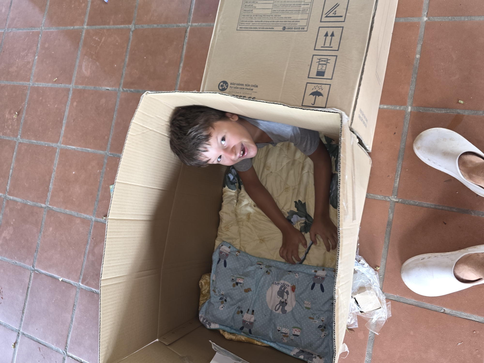 The image shows a young boy inside a large cardboard box. The box contains a folded blanket and a pillow with a cartoon print. The boy is looking up at the camera and smiling, with his head and upper body visible through the opening of the box. A person is seen on the right side of the frame wearing white footwear. The scene appears to be set on a tiled floor, possibly a patio. Text printed on the box includes "BẢO HÀNH, SỬA CHỮA TU VẤN SẢN PHẨM CUSTOMER SERVICE 1900 5555 66" indicating possible customer service details. It is in a location of Mũi Né, Vietnam, indicated by the language used on the box.