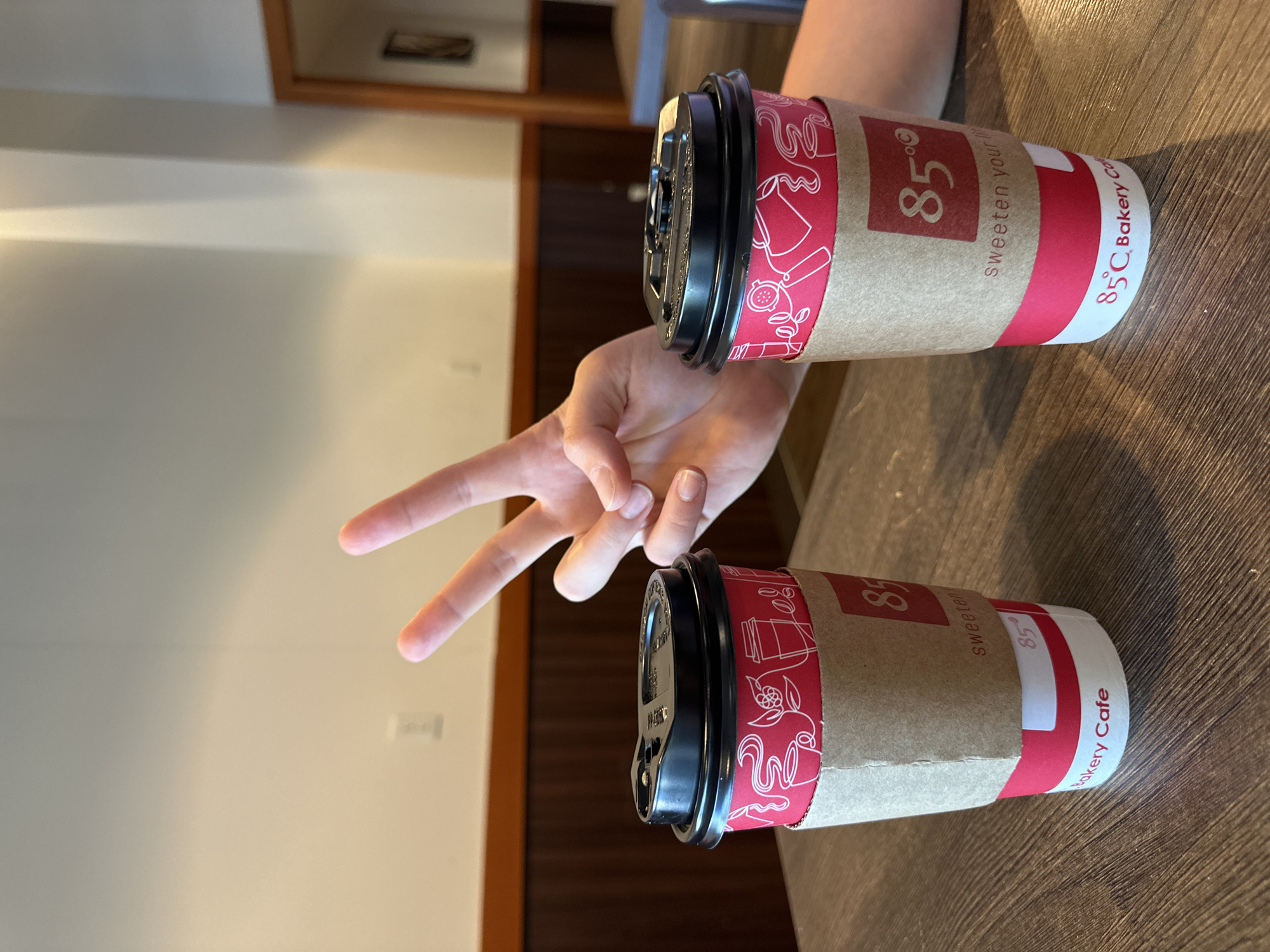 An over-the-shoulder medium shot captures two red paper coffee cups with brown cardboard sleeves resting on a wooden table. A left hand with three fingers extended in a peace sign gesture is visible in the upper right portion of the frame, partially obscuring the cups. The cups are branded with "85°C Bakery Cafe" and feature a white line-art design of coffee cups and coffee beans. The background shows a light-colored wall with brown wood paneling and a glimpse of a framed picture in a mirror on the far right. The setting appears to be an indoor cafe in Lynnwood, Washington, United States of America.