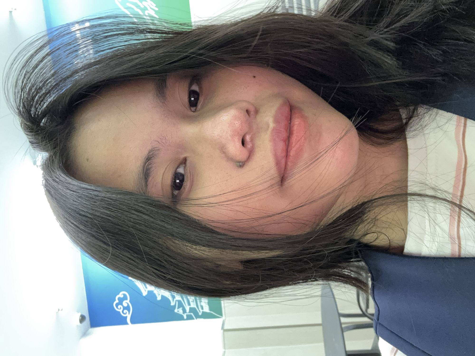 The image shows a close-up selfie of a young woman with long, dark hair. She is wearing a dark blue jacket over a white shirt with thin pink stripes. The background features a wall with a blue and green mural depicting clouds and traditional Asian architecture, and a white, pleated partition. A dark table and chairs are partially visible on the left. The scene appears to be indoors, possibly in a cafe or public space in East Jakarta, Indonesia, given the style of the mural. A security camera is visible on the ceiling in the upper left corner.
