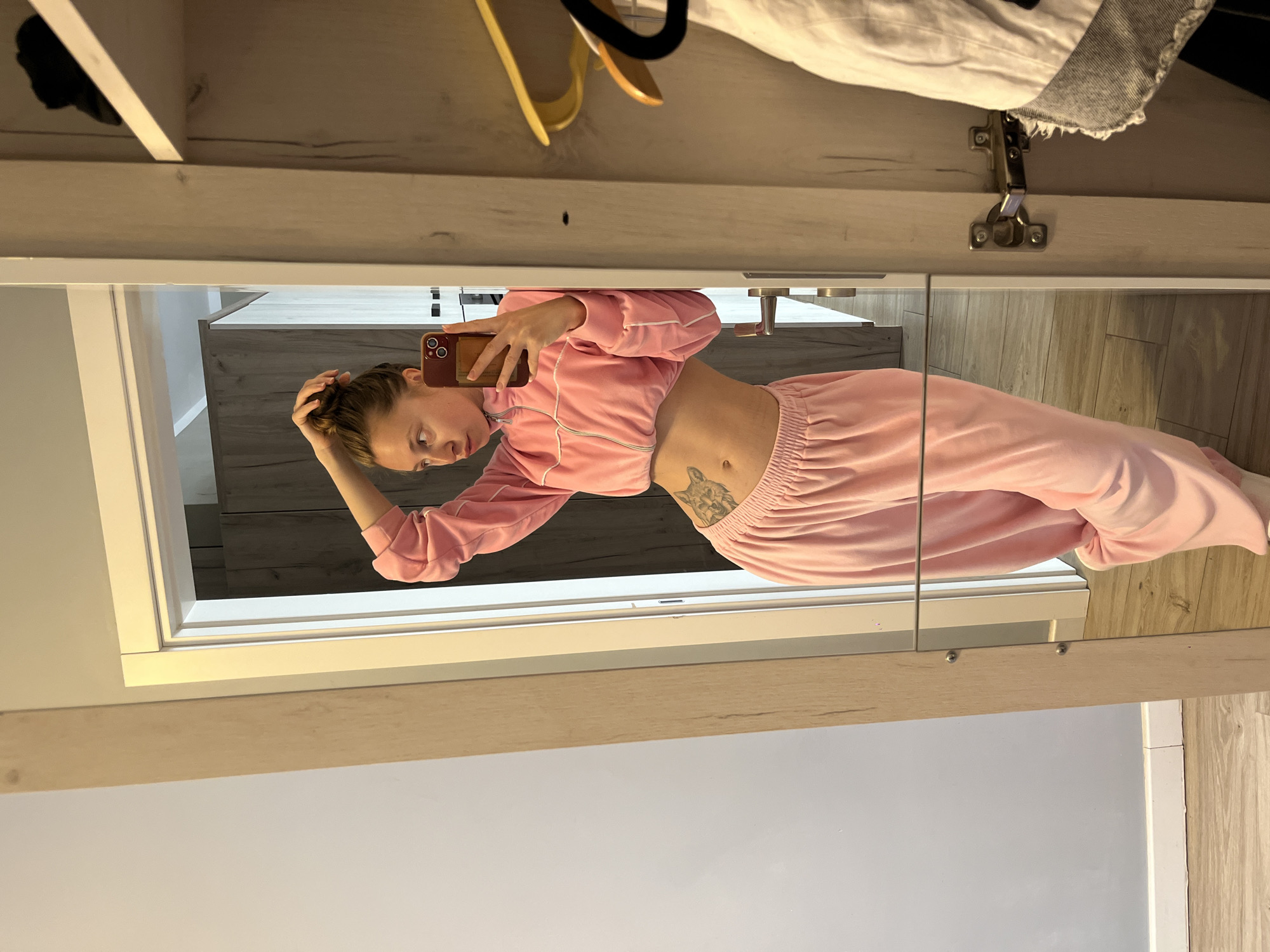 A woman is taking a selfie in a mirrored wardrobe. She is wearing a pink velour tracksuit consisting of a cropped jacket and pants. A tattoo is visible on her abdomen. The wardrobe contains clothes hangers and other items. The background of the wardrobe reflects part of a room. The scene appears to be indoors, possibly in Rafailovići, Montenegro.