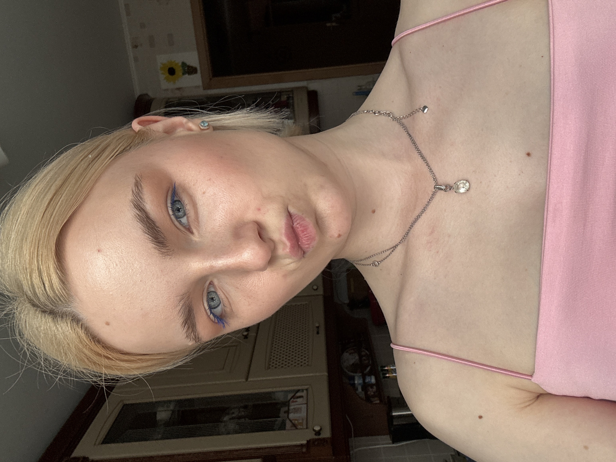 A young woman with blonde hair pulled back and blue eyeliner on her eyelids poses for a selfie. She is wearing a pink tank top and a silver necklace with a clear pendant. Her lips are pursed as she looks at the camera. The background shows a kitchen with wooden cabinets and a sunflower painting on the wall. The lighting suggests it is daytime. The image is taken from a close-up, chest-up perspective.