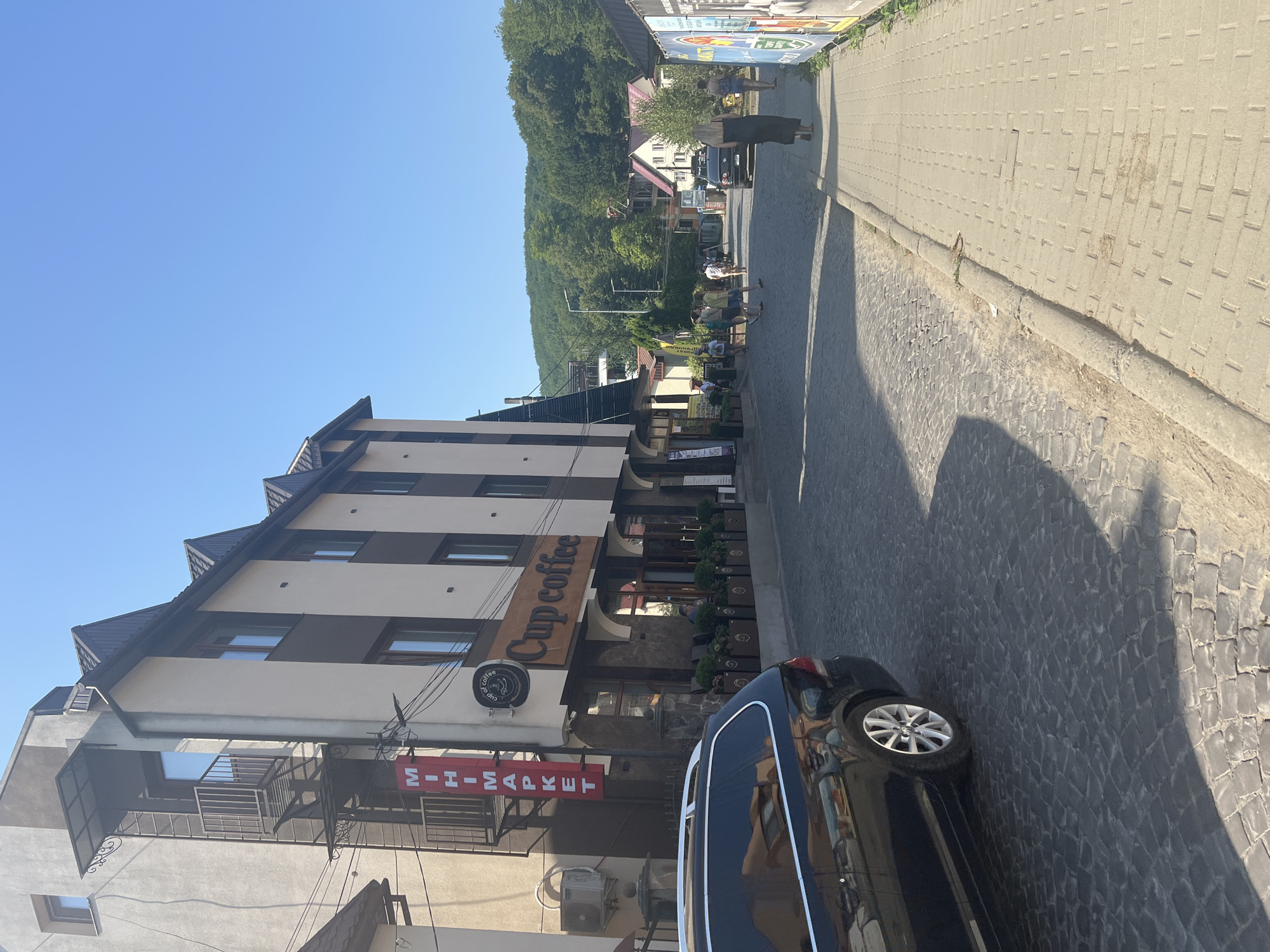 A sunny day in Сонячне, Ukraine, features a cobblestone street lined with buildings and a paved sidewalk. On the left, a multi-story building with a brown and white facade houses a "Cup Coffee" shop. A red vertical sign above it reads "MIHIM MARKET." In the foreground on the left, a black station wagon is parked. The street is populated by several people walking in various directions. In the background, lush green hills rise under a clear blue sky. The scene depicts a typical day in a Ukrainian town with a mix of commercial establishments and natural scenery.