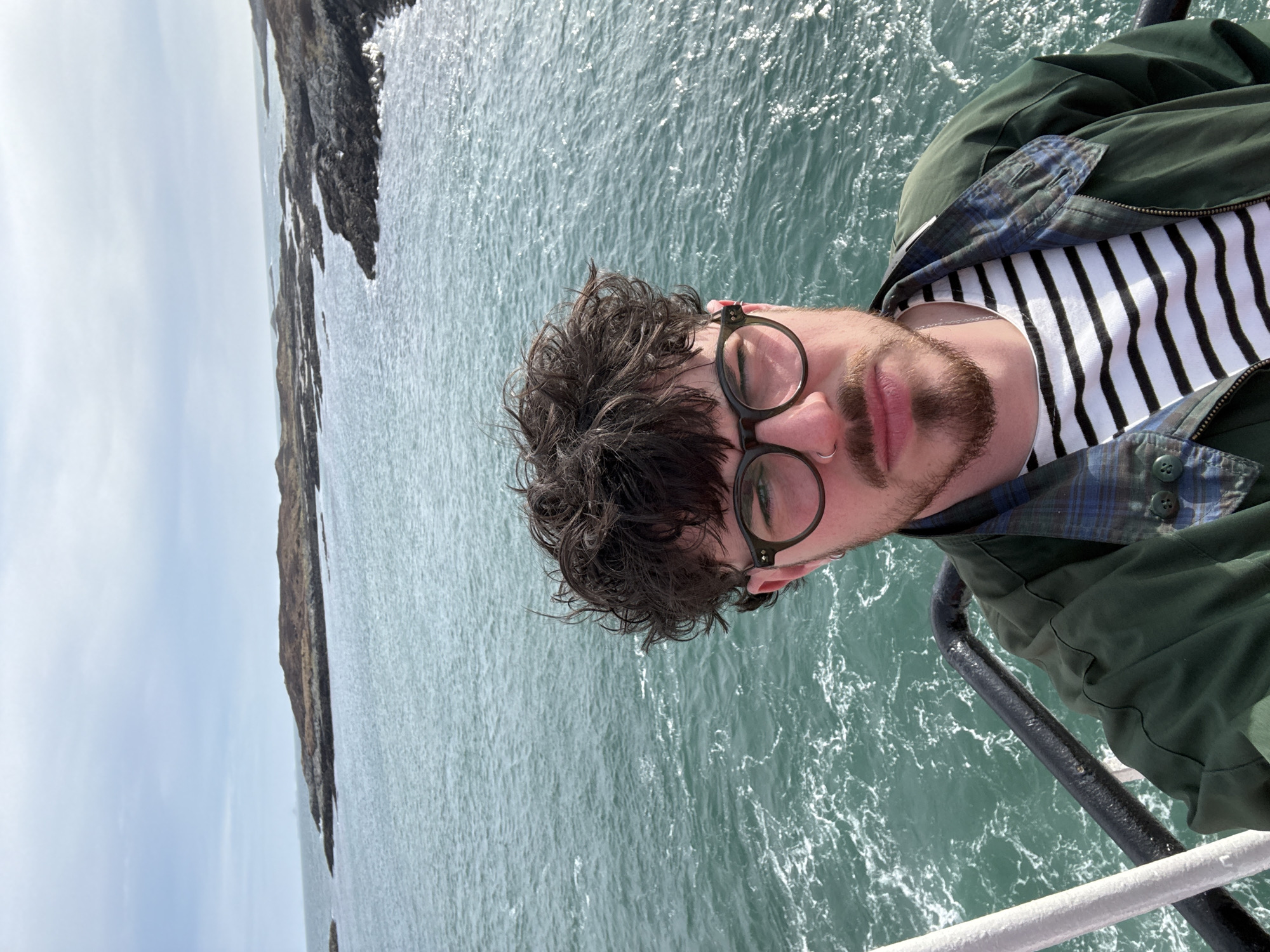 A medium shot depicts a man wearing glasses, a striped shirt, and a green jacket standing in front of a body of water. Behind him is a rocky coastline under a cloudy sky. The man is leaning against a black railing. The location is unknown.