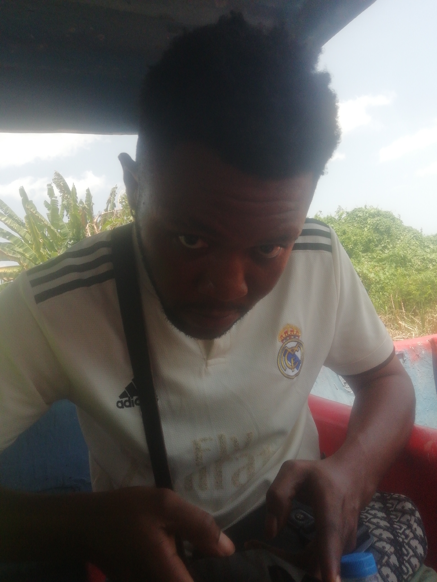 A close-up photo shows a dark-skinned man wearing a white Real Madrid football jersey with black Adidas stripes on the shoulders. A black strap is slung across his chest. The background reveals green vegetation, possibly banana trees and shrubs, suggesting a tropical environment. The image appears to have been taken outdoors under daylight conditions, with glimpses of a cloudy sky. The man seems to be inside a structure with a red interior. The location might be the city of Ikorodu, Nigeria, considering the user's prompt.