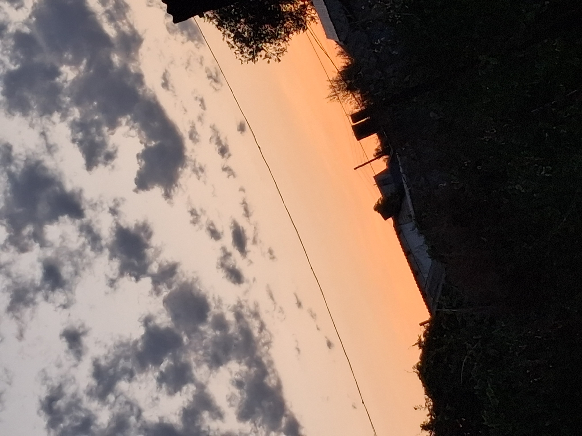 The image depicts a twilight sky over a residential area in Semey, Kazakhstan. The sky is a gradient of pale orange near the horizon, transitioning to a lighter, hazy gray-blue towards the top. Dark, scattered clouds are visible against the lighter sky. A thin power line stretches diagonally across the frame from the upper left to the lower right. In the foreground and lower portion of the image, the silhouettes of buildings, trees, and dense vegetation are visible against the fading light. The structures appear to be low-rise buildings, possibly houses, with dark roofs. The overall impression is that of a rural or suburban landscape at dusk.