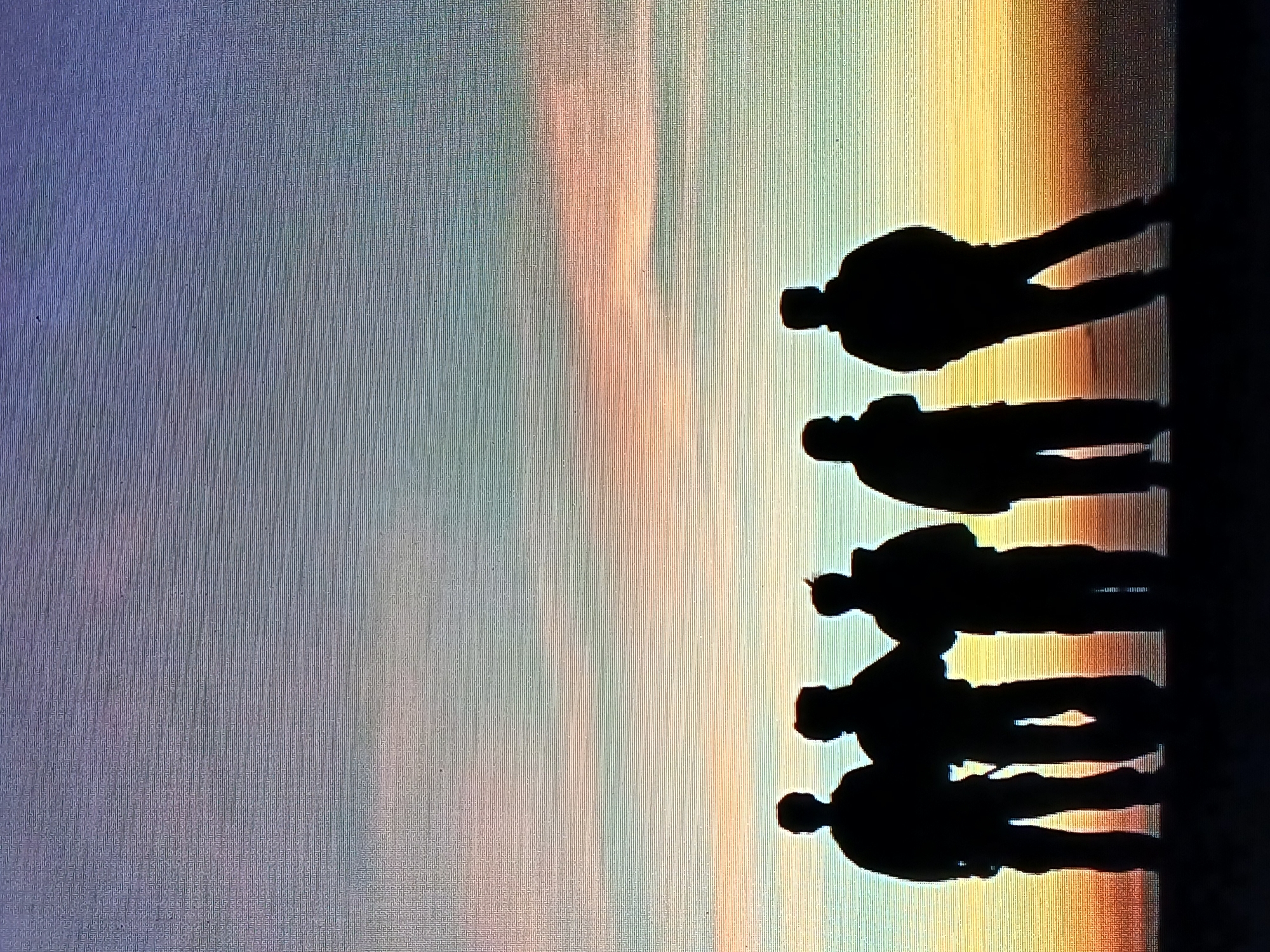 The image depicts five silhouetted figures standing in a line against a colorful sky, transitioning from purple and blue to orange and yellow. The location is likely Juan Griego, Venezuela. The figures are all facing in the same direction. The ground or surface they are standing on appears to be dark and flat.