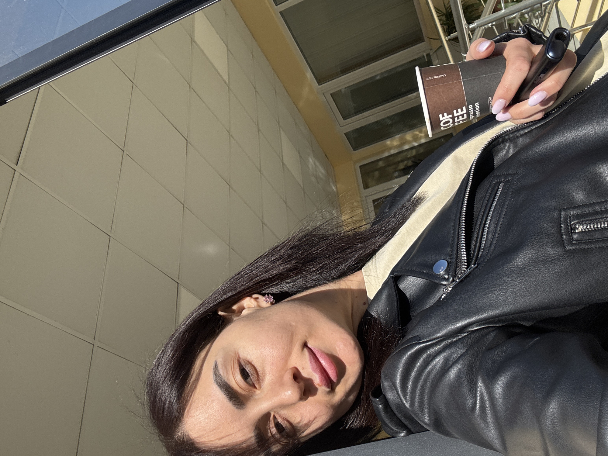 A person, likely a woman, is pictured in a selfie-style photograph. She has dark hair and is wearing a black leather jacket. In her hand, she holds a coffee cup and what appears to be an electronic cigarette or vaping device. The coffee cup has the words "Coffee Espresso Emotion" printed on it. The background appears to be the exterior of a building with a tile or panel facade. Based on visual characteristics, it is likely that the location is in Tbilisi, Georgia.
