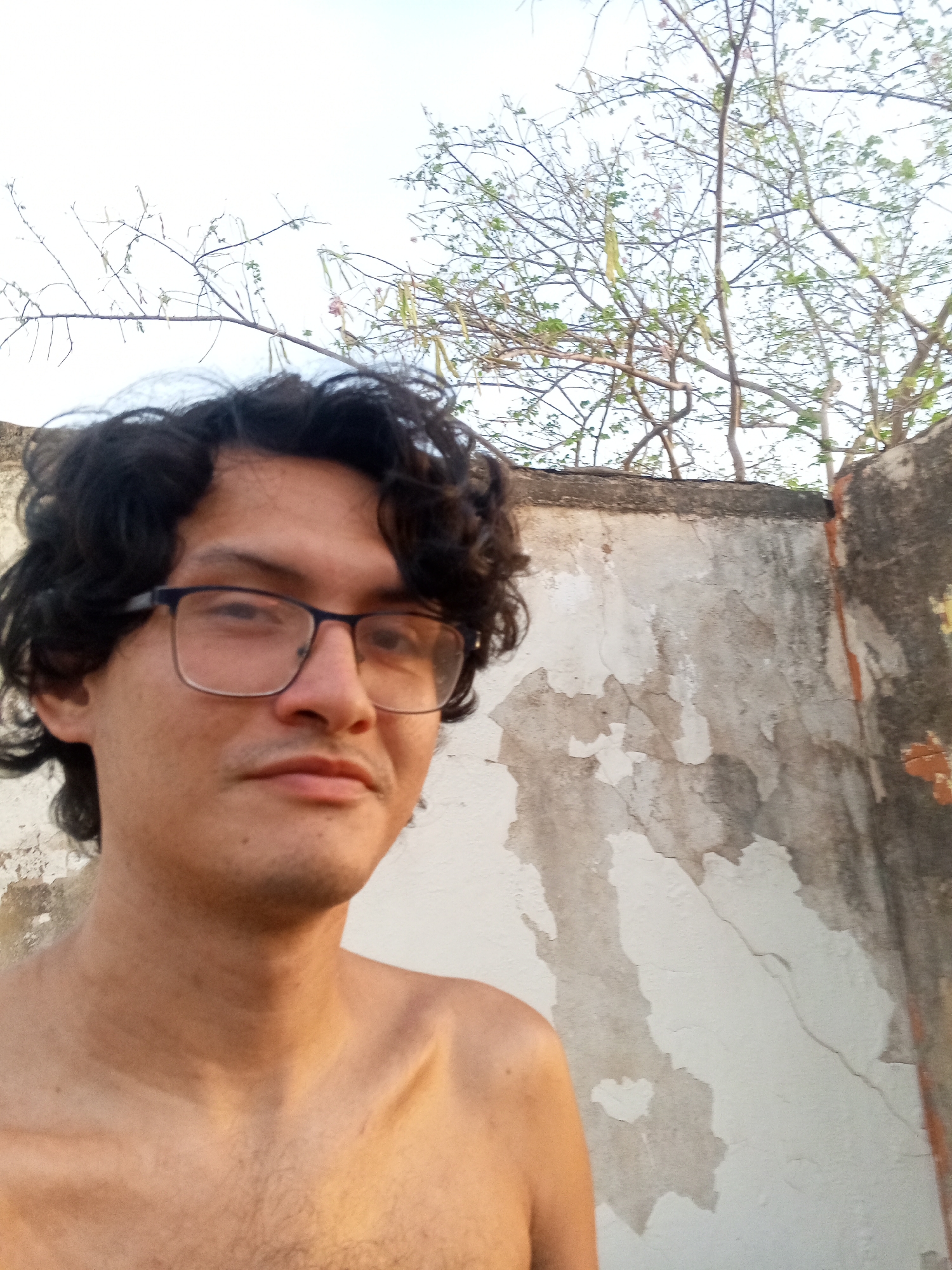 A shirtless man with dark, curly hair and glasses is taking a selfie outdoors. He is positioned in front of a dilapidated wall with peeling paint and a tree with sparse foliage visible above. The location is Juan Griego, Venezuela, judging by local architecture and vegetation.