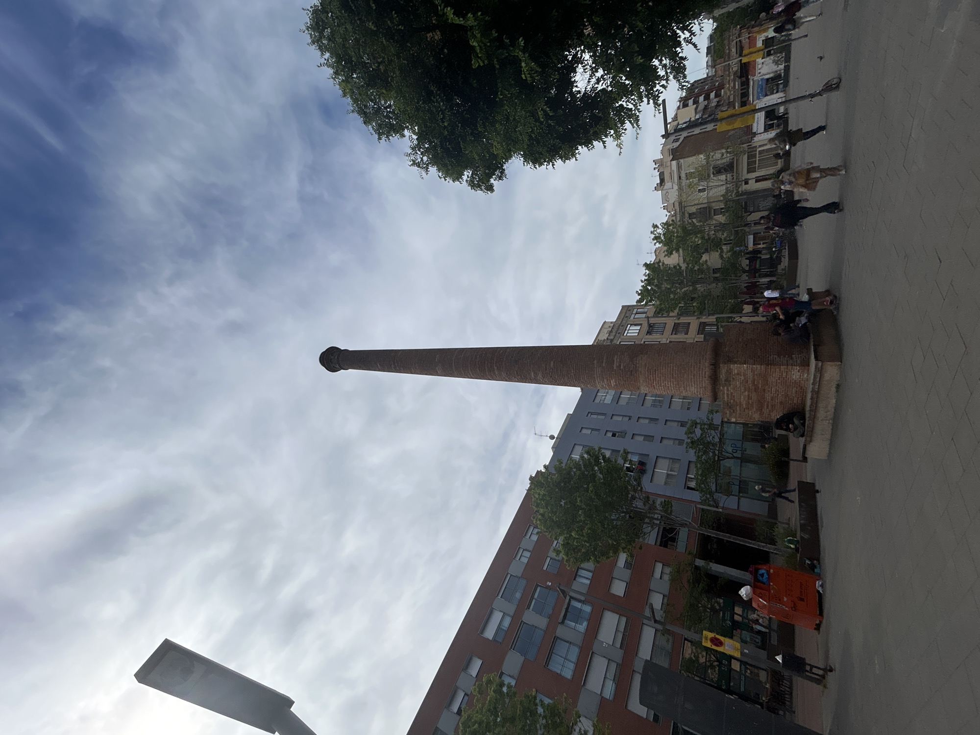 The image shows a tall brick smokestack, likely the Espanya Industrial Park Chimney in Barcelona, Spain. The sky is cloudy, with trees visible above. Modern buildings of various colors stand adjacent to the stack. People walk along a paved area near the base of the smokestack, some sitting on a low brick wall. A bright orange trash container is visible in the foreground. A street lamp is also located in the foreground and partially visible.