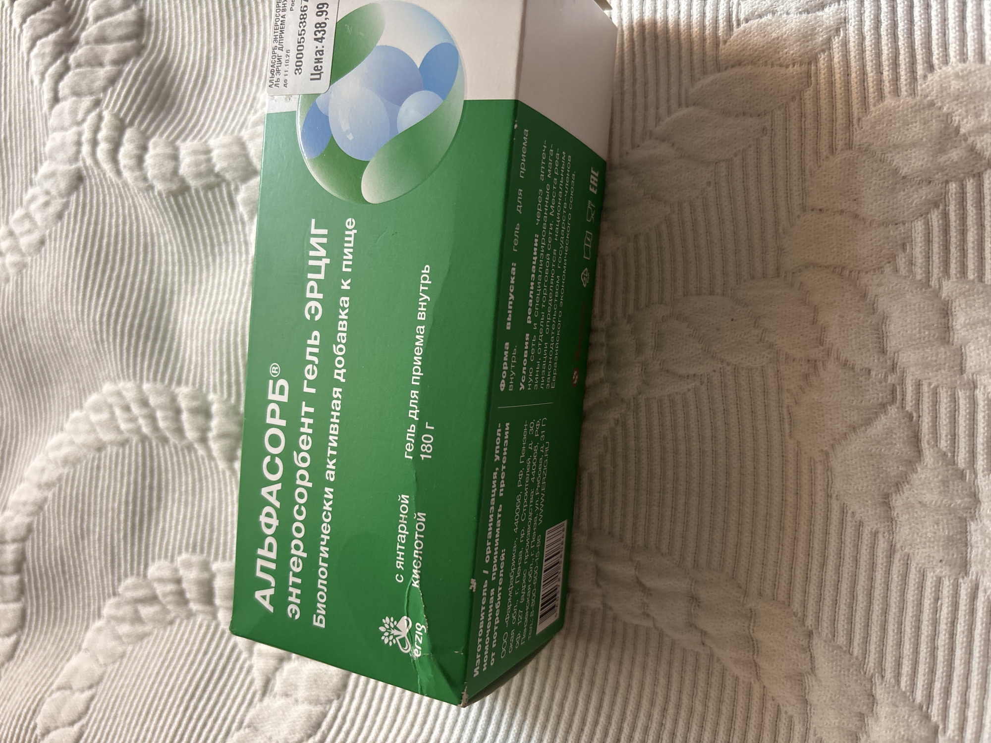 The image shows a close-up of a green box with Russian text. The box appears to be for a health product called "АЛЬФАСОРБ® энтеросорбент гель ЭРЦИГ" (Alphasorb® enterosorbent gel ERZIG). The text indicates it is a biologically active dietary supplement. There is also a price tag on the box that reads "Цена: 438,99" (Price: 438.99). The box is resting on a white, textured fabric, likely a blanket or bedspread. The background is out of focus and shows a repeating pattern of raised lines and diamond shapes, typical of knitted fabric. There is no indication of the city of Chernogorsk, Russia, or any specific location within it. The image is a still shot and does not depict any actions or interactions.