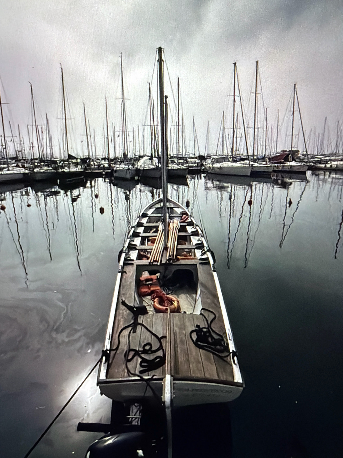 The image shows a fleet of sailboats docked in a harbor, with one small boat in the foreground. The sky is overcast and the water is calm, reflecting the sailboats. The foreground boat is made of wood and has a checkered pattern on the inside. There are life vests and other equipment on board. The background shows many other sailboats with tall masts. The location is identified as Kafr Yasif, Israel.