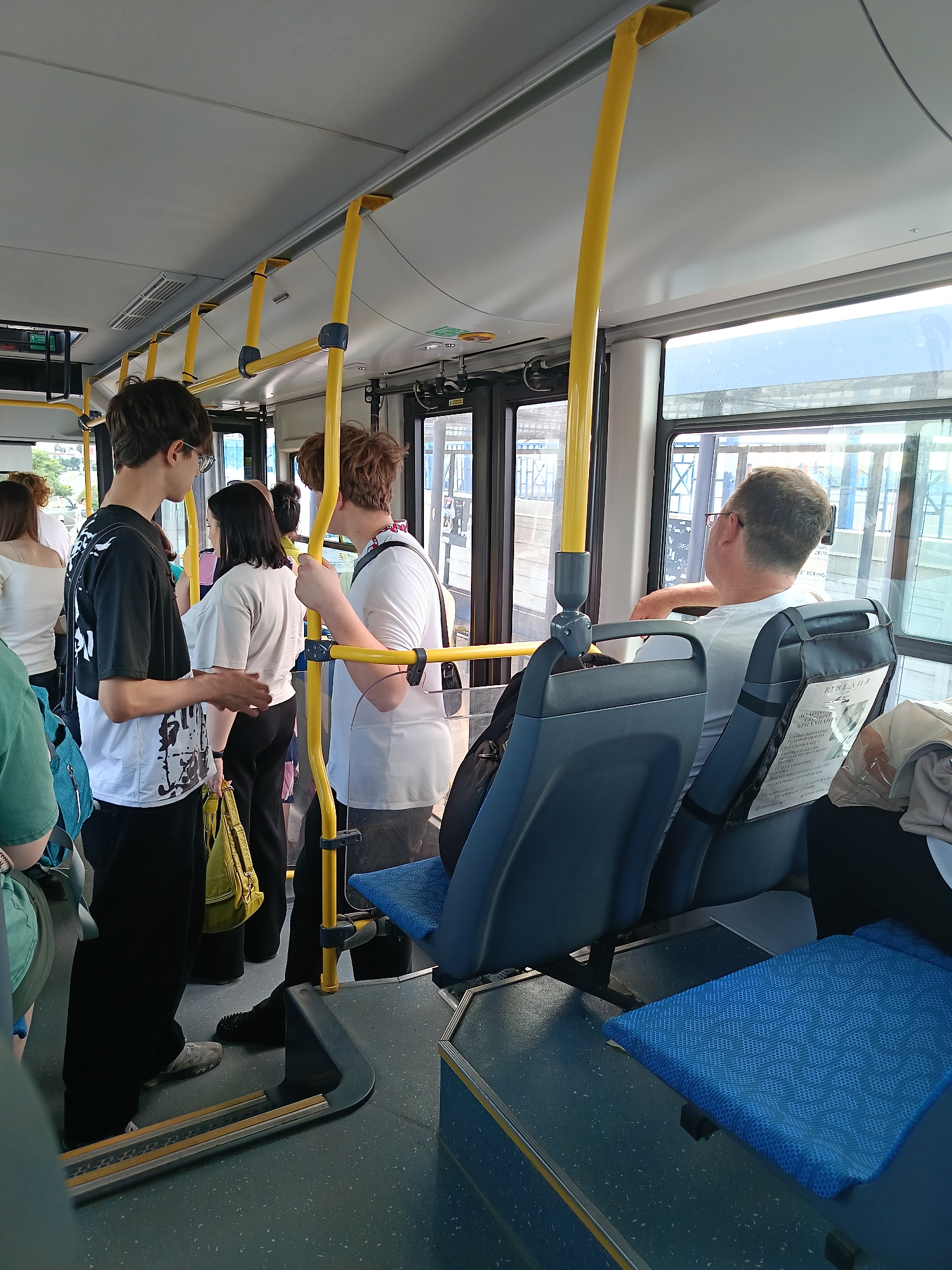 The image depicts the interior of a public bus, likely in Gubkin, Russia, given the Cyrillic text on the seatback. Several passengers are visible, some standing and others seated. In the foreground, a young person wearing a black and white t-shirt and black pants is standing, facing to the right and gesturing with their hands. Beside them, a young person with dark hair and a light-colored t-shirt is also standing. Further back, a person with reddish-brown hair wearing a white t-shirt is standing near a yellow pole. In the seats, a man wearing glasses and a white t-shirt is looking out the window. The bus has blue seats with yellow vertical poles and overhead handrails. The floor appears to be a speckled grey material. A blue cushion is placed on a seat in the lower right of the frame. The lighting suggests it is daytime, with natural light coming through the windows.