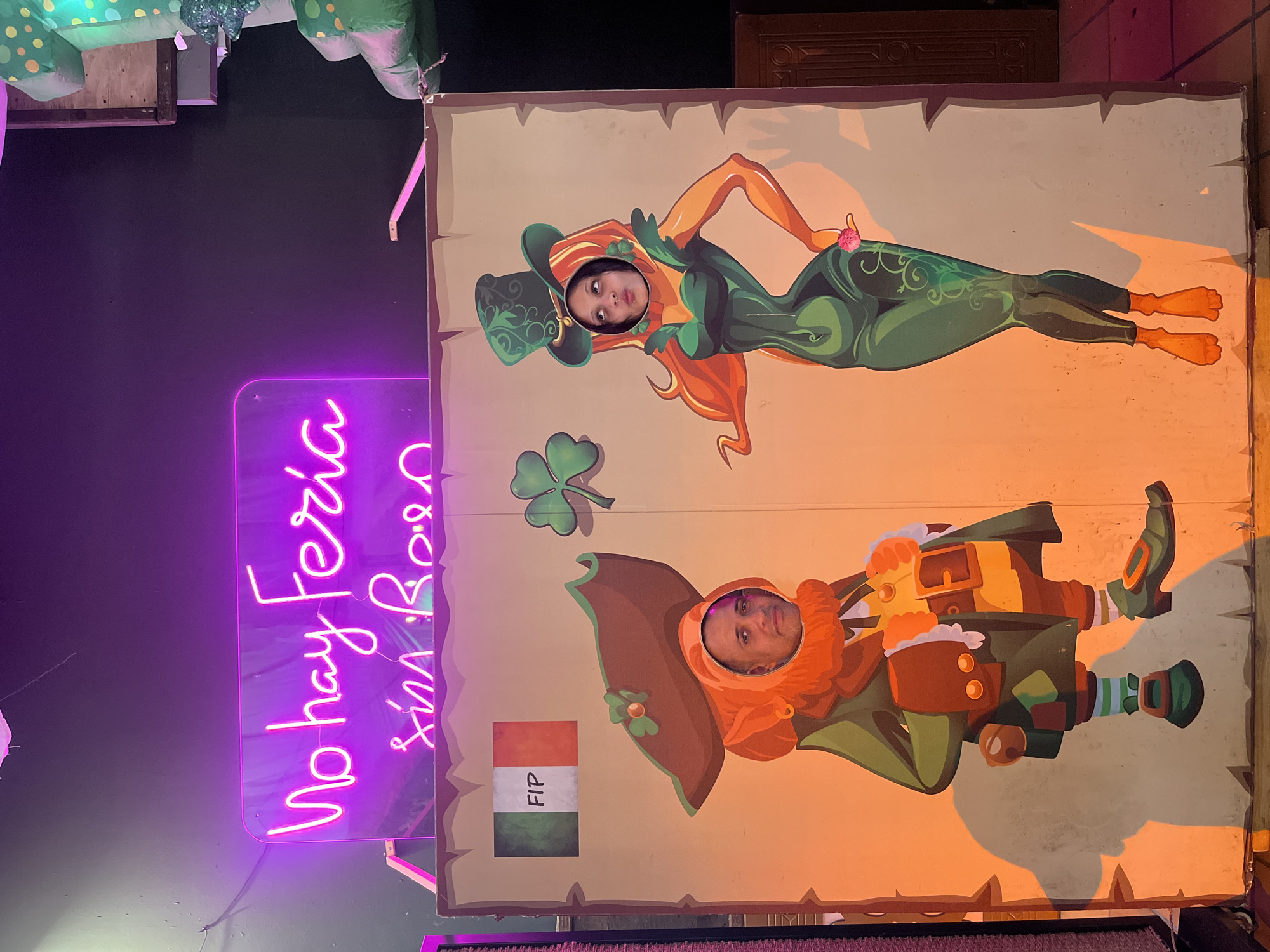 This media file captures an indoor scene featuring two people posing for a photo using a themed cutout board, likely at an event or venue in Fuengirola, Spain.
The central focus is a large, upright photo board featuring two cartoon characters: a female figure resembling a seductive leprechauness or nature spirit with long red hair, a green hat, and a leafy green suit, and a male leprechaun character with an orange beard, a brown hat, and green and brown attire, holding a pot of gold. A green shamrock is depicted between the two figures on the board. A woman's head is visible through the cutout for the female character, and a man's head through the cutout for the male leprechaun. Both individuals look towards the camera with neutral expressions, participating in the playful photo opportunity.
To the left of the board, a bright pink/purple neon sign glows in the dark background, displaying partially visible text that reads "No hay Feria sin Ro..." (suggesting "No hay Feria sin Rumba" or "There is no fair without Rumba/Party"). On the photo board, below the male character's head, there is a small flag with horizontal stripes of red, white, and green, bearing the letters "FIP" vertically on the white stripe; this flag resembles the flag of Hungary. In the upper left corner, out of focus, are green, polka-dotted decorative items, possibly inflatable.
The dark surroundings and neon lighting indicate an evening or indoor setting, suggesting a festive or entertainment event. The leprechaun and shamrock motifs hint at a possible St. Patrick's Day celebration or a general Irish-themed event.