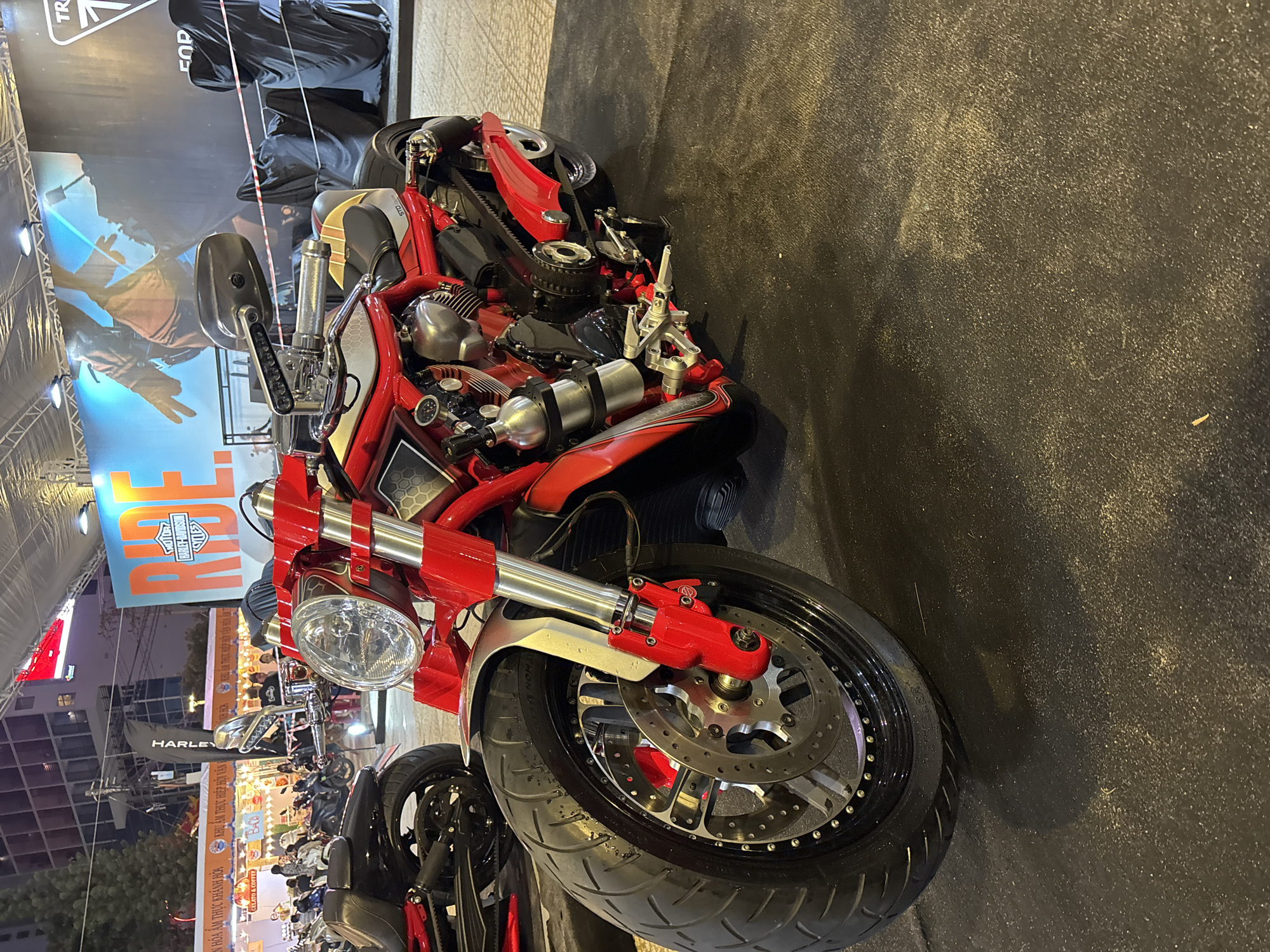 The image captures a close-up view of a red and silver motorcycle, potentially at an event or exhibition. The front wheel is prominently featured, with a clear view of the tire and disc brake. In the background, there is an outdoor setting, possibly in the city of Nha Trang Ward, Vietnam, where a sign written in Vietnamese can be seen. A Harley-Davidson sign and Triumph advertisement are also visible in the background, suggesting that the event is related to motorcycles. The street appears busy with people, although they are slightly blurred, indicating movement. The presence of multiple motorcycles suggests that this may be a motorcycle show or gathering.