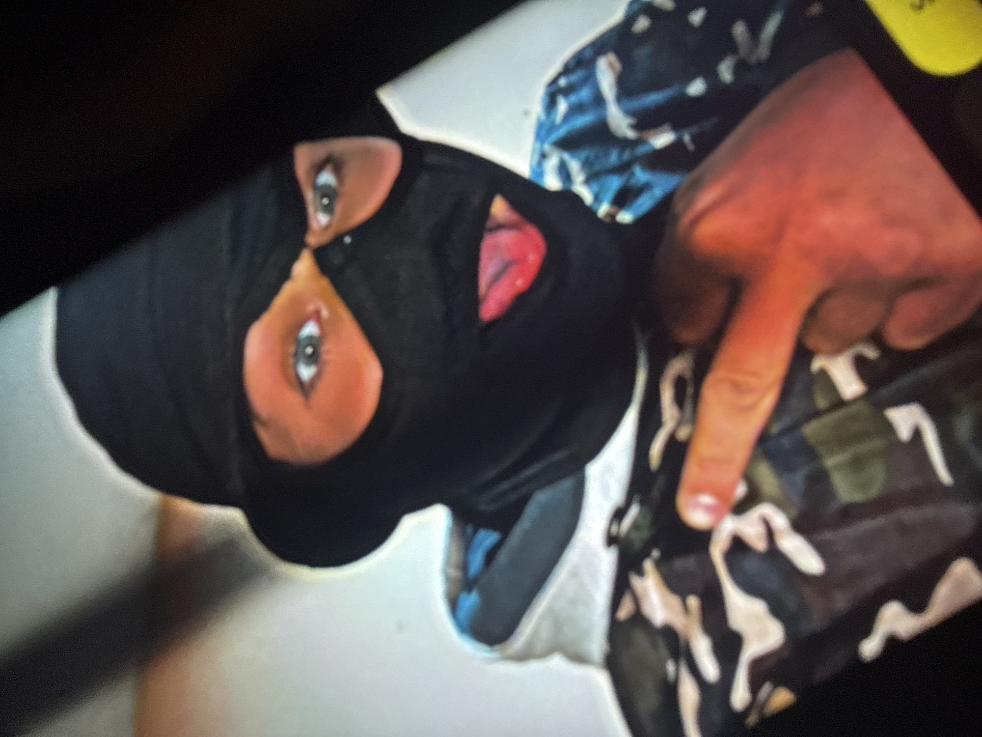 A person is wearing a black balaclava and a black hat. They are sticking their tongue out and wearing a camouflage jacket. Their right index finger is raised, pointing towards the viewer. The background is blurred and appears to be indoors. There is no discernible location in the city of Sofia, Bulgaria.