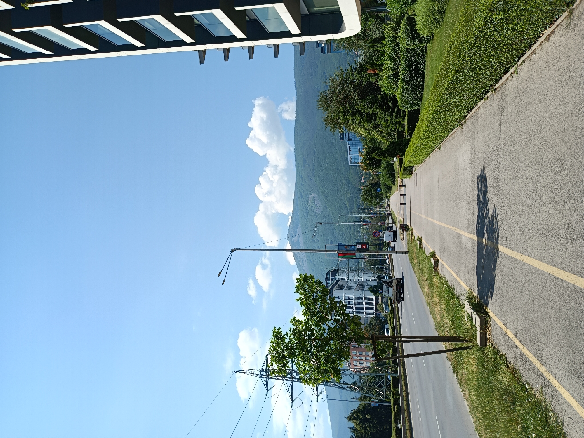 The image depicts a street scene in Sofia, Bulgaria, on a clear day. The perspective is from ground level, looking towards a road that curves to the right, bordered by a grassy median and meticulously manicured hedges. In the distance, a large, tree-covered mountain dominates the skyline, partially obscured by fluffy white clouds.
On the left side of the road, a modern building with a distinctive facade of dark panels and glass windows is partially visible. Across the street, a series of contemporary buildings with glass exteriors are nestled among lush green trees. A tall transmission tower with a network of wires stands prominently in the foreground, with a vibrant green tree growing in front of it. Streetlights and traffic signals are also visible along the road.
The road itself is asphalt, marked with yellow lines and a white line, suggesting a dedicated lane for bicycles or pedestrians. The time of day appears to be daytime, given the bright sunlight casting shadows on the road. The overall impression is one of an urban environment with significant green spaces and a backdrop of natural landscape. There is no visible text in the image.