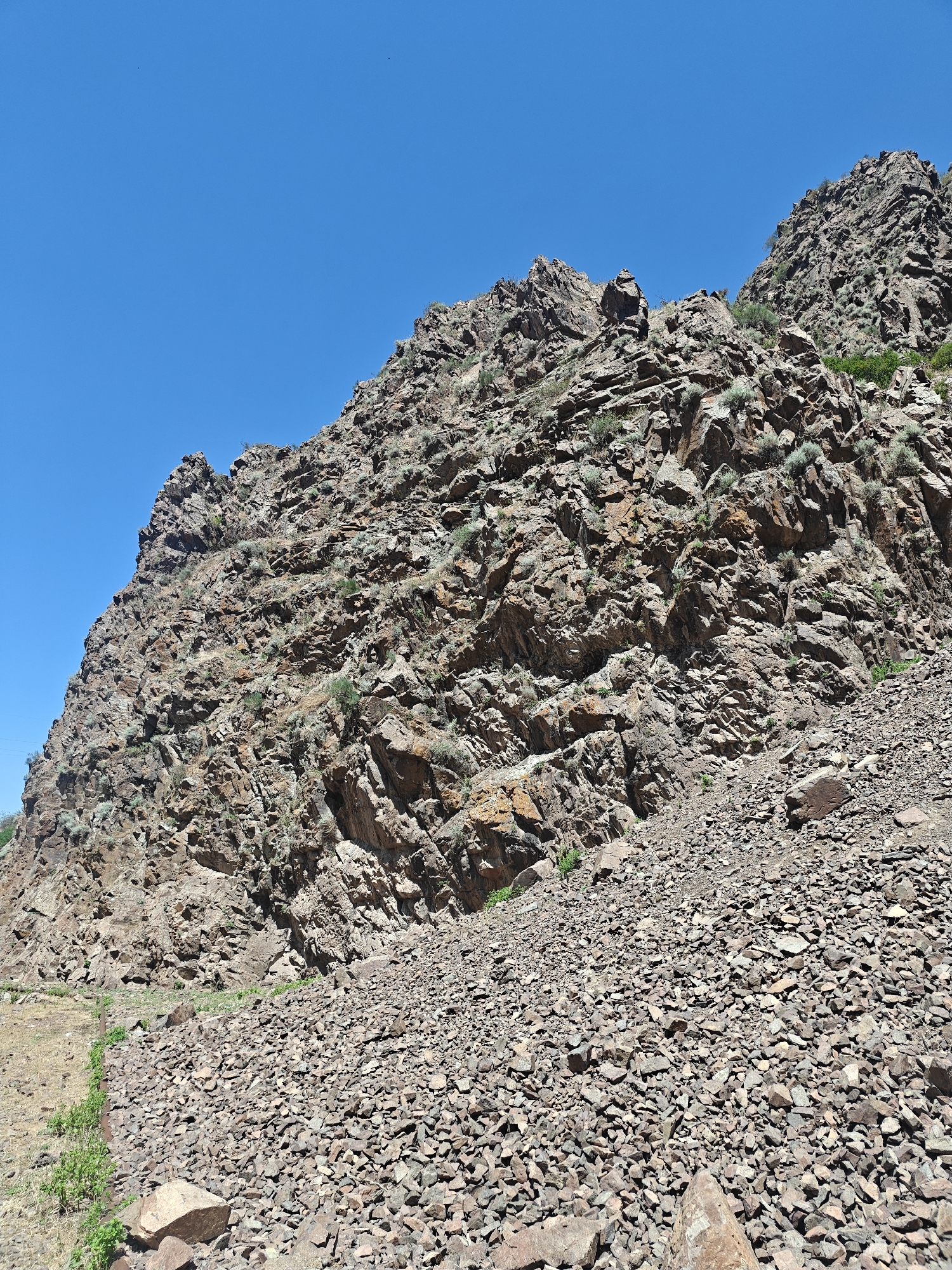 A bright, sunny day in Малиновка, Kyrgyzstan, reveals a steep, rocky mountainside under a clear blue sky. The terrain is rugged and uneven, covered with numerous loose rocks and stones of varying sizes. Sparse, dry vegetation clings to the rocky slopes, adding touches of green to the otherwise earthy tones of the mountain. A narrow, dirt path is visible along the left side of the image, winding its way up the lower part of the mountain. The overall scene depicts a natural, arid landscape with no human presence or activity evident.