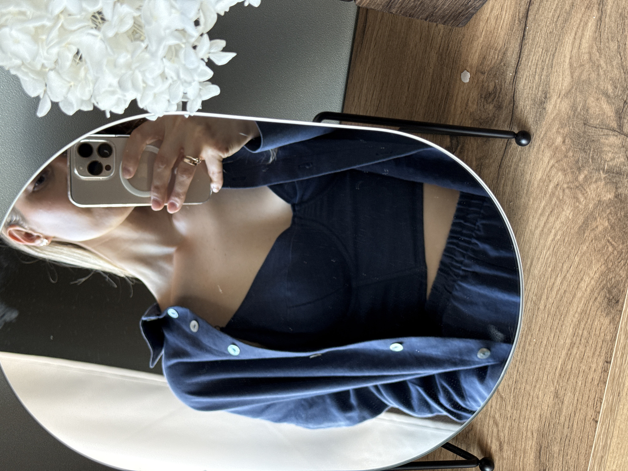 The image is a mirror selfie taken indoors. A woman, partially visible, is holding a smartphone to take the photo. She is wearing a dark blue shirt and a dark top. The background includes a white floral arrangement, a gray wall or surface, and a wooden surface. The location details of Chaikovsky, Russia, are not discernible from the visual information provided in the image.