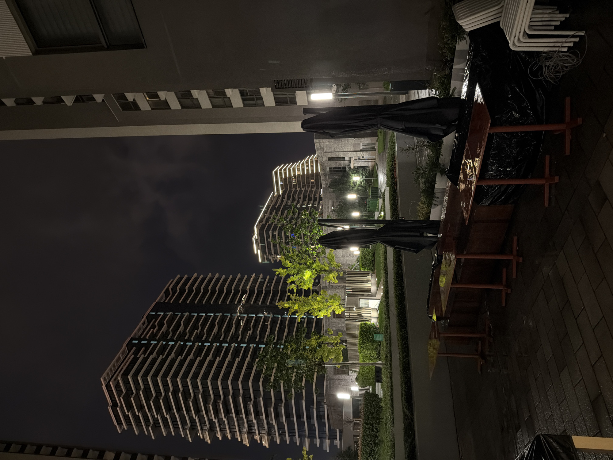 This is a night scene at the waterfront in Belgrade, Serbia, likely near the Savamala district, focusing on a cafe or restaurant outdoor seating area. In the foreground, several tall, narrow tables with orange metal bases are covered in a black tarp, suggesting they are closed for the season or due to weather. Two folded patio umbrellas, also covered in black fabric, stand nearby. Stacked white chairs are visible to the right. The ground is wet, indicating recent rain.
Behind the seating area, a well-maintained lawn and landscaping with illuminated trees and bushes are bordered by a low wall. Several modern, multi-story buildings dominate the background. The most prominent is a tall, contemporary residential building with horizontal balconies and a facade that reflects light. Further in the distance, two other similar buildings are illuminated with decorative lights along their upper floors. The sky is dark and overcast. The overall impression is one of an urban, modern setting at night, with outdoor amenities temporarily out of service.