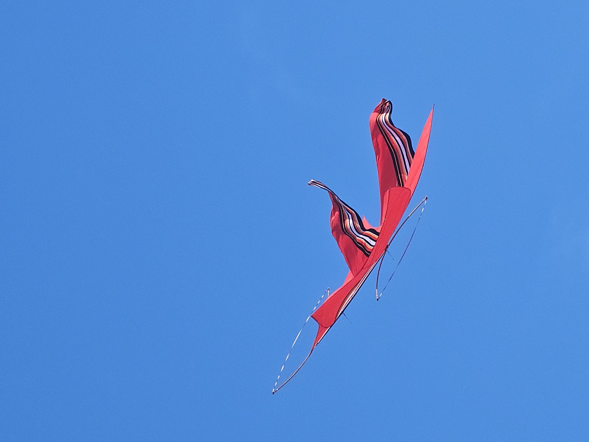 A red kite with black, white and orange stripes flies in a clear blue sky. The photo was taken during the Bali Kite Festival in Denpasar, Indonesia.