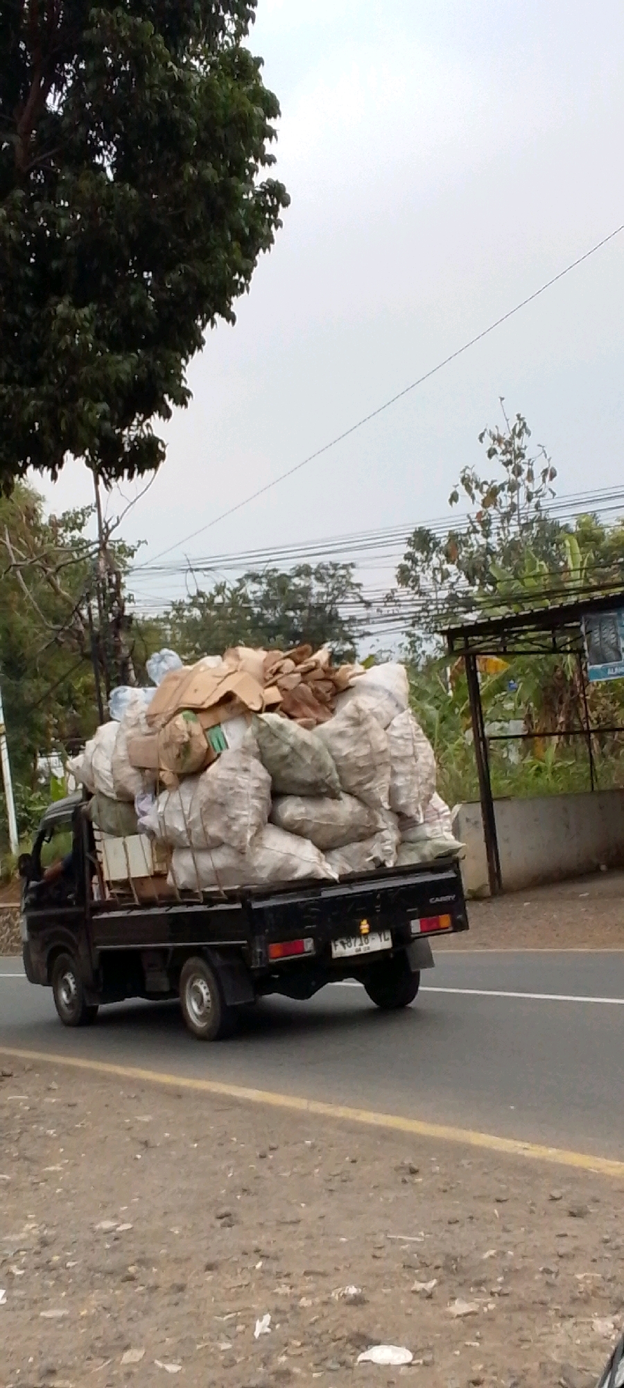 A dark-colored utility truck is driving on a paved road in Cilaku, Indonesia. The truck is heavily loaded with large sacks and cardboard boxes, which appear to contain recyclable materials or agricultural produce. The truck is moving from left to right in the frame, and its license plate is partially visible, reading "F 8718 YL". The surrounding environment includes trees, power lines, and a cloudy sky. The road has a yellow line marking its edge. The ground next to the road is dirt and appears to be littered with some debris. A man is visible in the driver's seat of the truck.