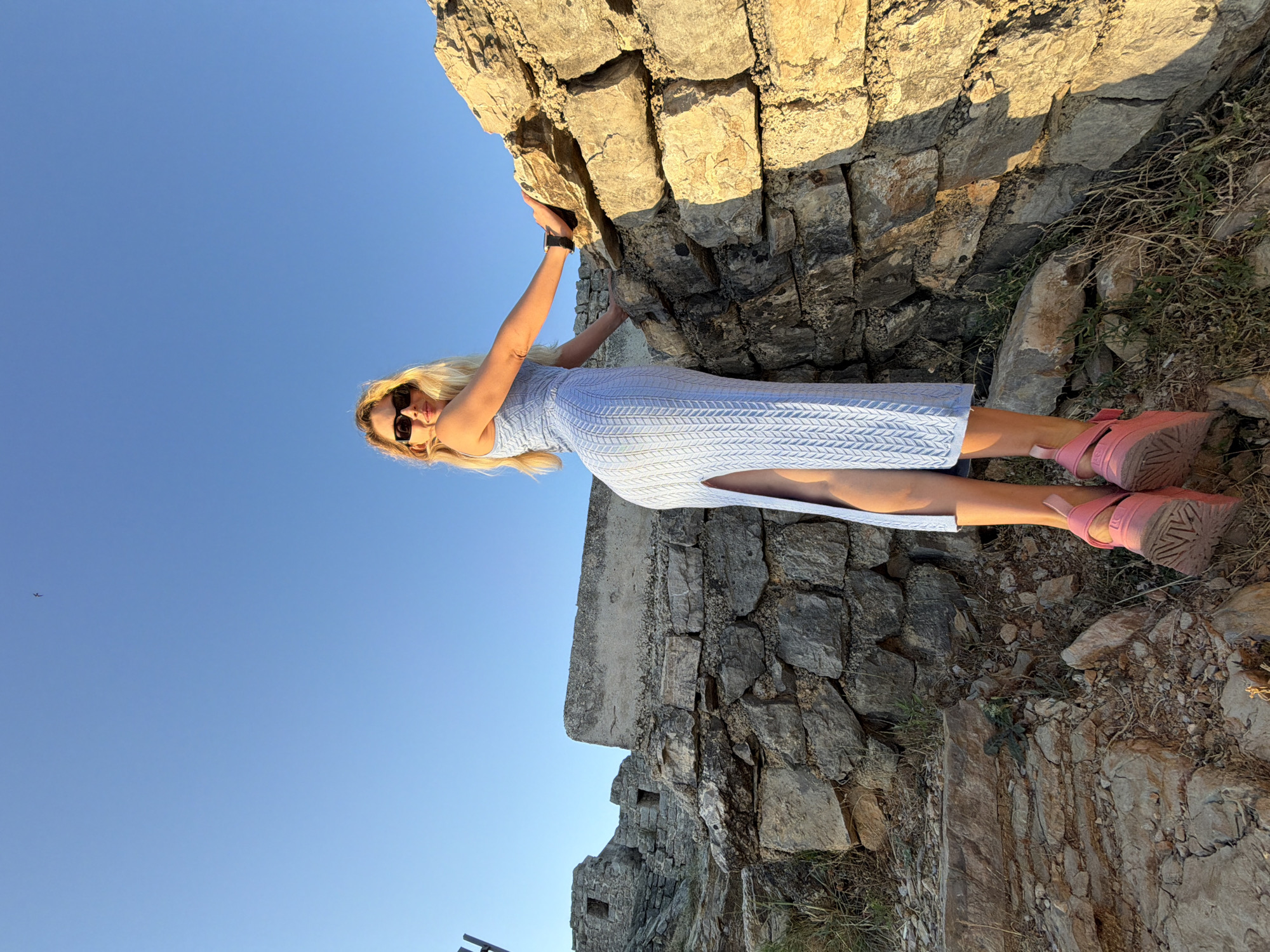 This image depicts a woman posing in an outdoor setting characterized by ancient stone ruins, identified as being in Budva, Montenegro. The woman, centrally positioned and slightly angled, is a blonde individual wearing black rectangular sunglasses. She is dressed in a light blue, sleeveless, form-fitting knit midi-dress featuring a distinct patterned texture and a high slit on her left side. Her footwear consists of pink platform sandals with rugged soles. A dark wristwatch is visible on her left wrist. She is leaning with her right hand on a rough-hewn stone wall to her right, while her body is turned slightly to the right, and she is looking directly at the camera over her right shoulder.
The immediate background behind the woman consists of more sections of dry-stacked stone walls. Further into the background, to the left, are larger remnants of stone structures, possibly ancient buildings, with visible openings resembling windows. The ground beneath the woman's feet and surrounding the ruins is rocky, dry, and sparse with withered vegetation. The sky is clear and bright blue. The scene is illuminated by strong, warm sunlight, casting defined shadows and highlighting the texture of the stone. High in the upper right portion of the sky, a small, dark, bird-like object is discernable.