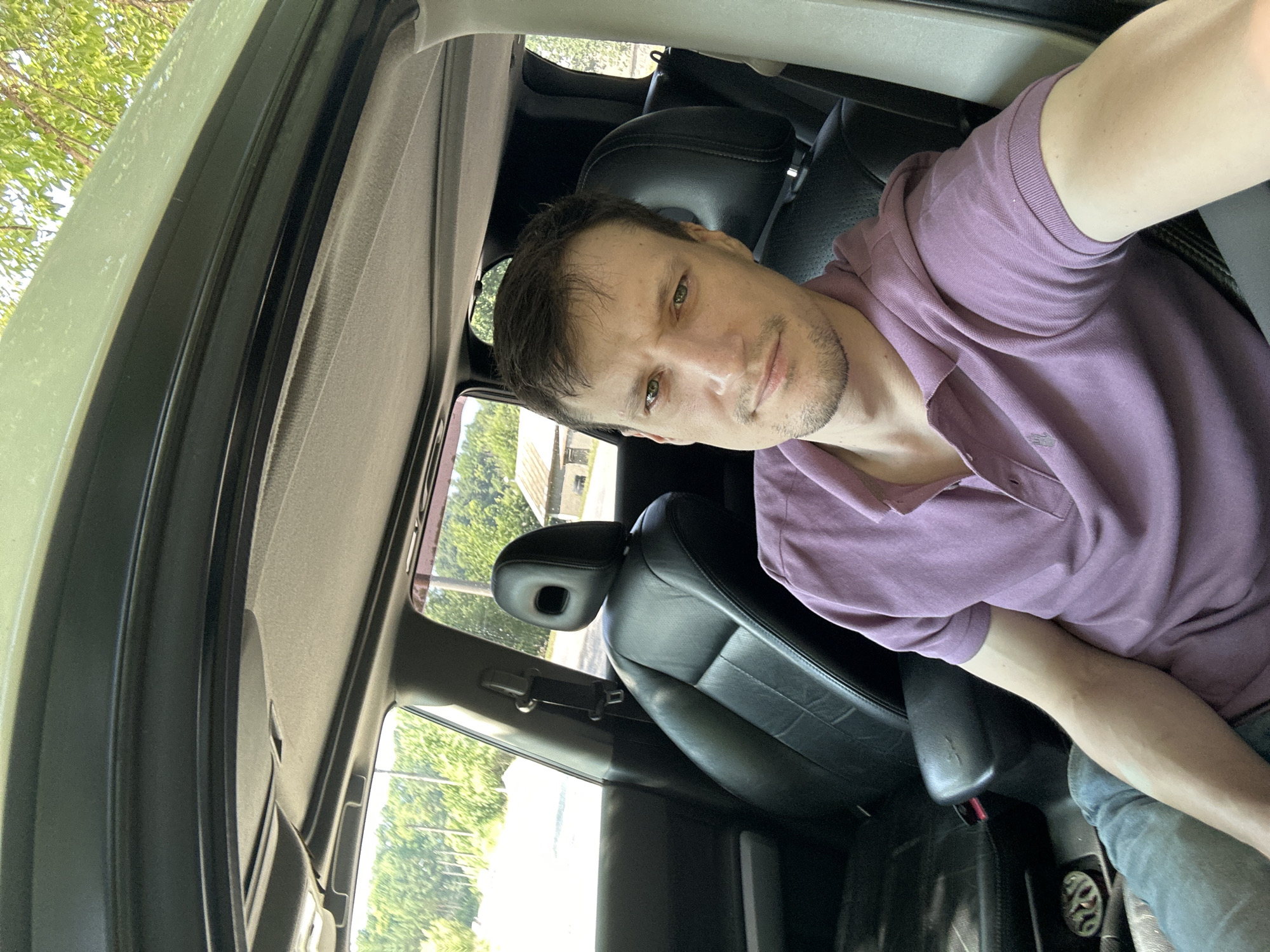 A selfie taken from inside a car shows a Caucasian male with light skin, dark brown hair, and green eyes. He has stubble on his face and is wearing a purple polo shirt. The interior of the car is visible, including black leather seats and a sunroof. Outside the car, a rural landscape with trees and a building can be seen. The overall lighting suggests it is daytime. The image is likely taken in Makiv, Ukraine, based on the provided context, however, no specific landmarks are identifiable within the image to confirm this location.
