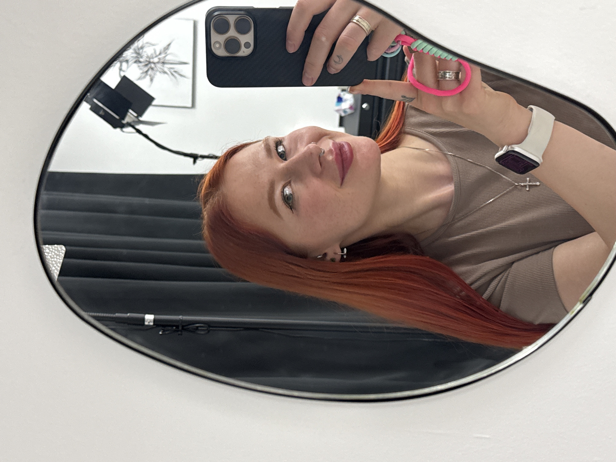 A red-haired woman is taking a selfie in a mirror. She is wearing a beige top and a silver necklace with a cross pendant. She has multiple rings on her fingers and a smartwatch on her wrist. Her phone, a black iPhone, is visible in the shot. The mirror reflects a room with a black curtain or backdrop, a lighting setup, and a piece of art on the wall. The location is not definitively identified as Rostov-on-Don, Russia, as there are no specific visual cues within the image to confirm this.