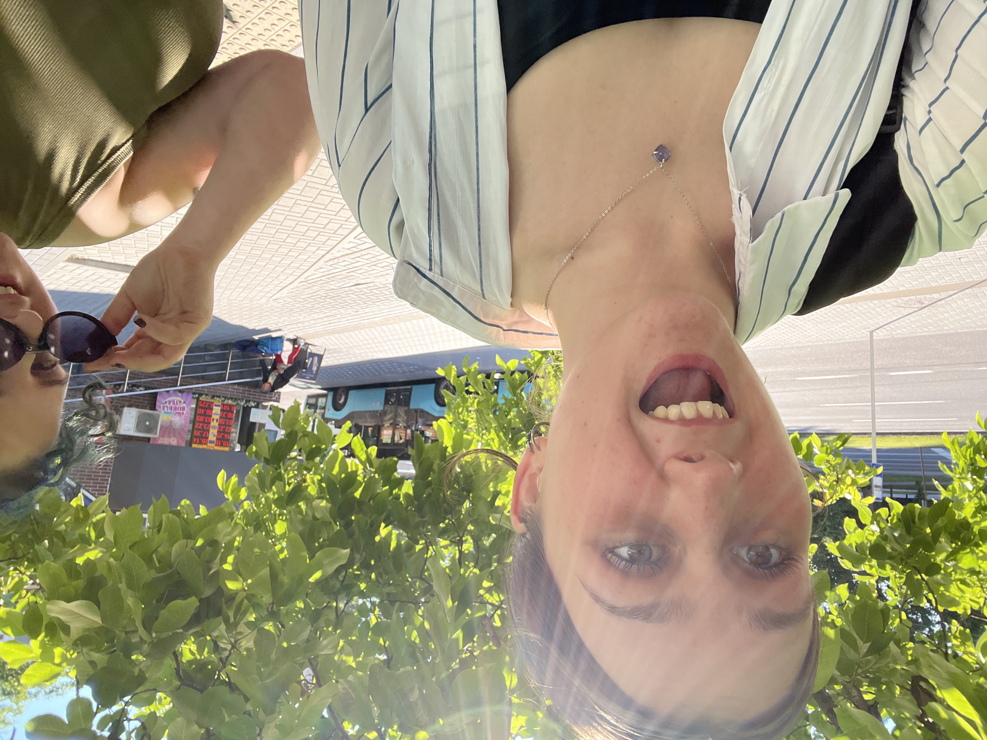 This image shows two women outdoors, possibly in a city. The woman in the foreground, on the left, is taking a selfie, with her mouth open in surprise or excitement. She has brown hair, light skin, and is wearing a white and blue striped open shirt over a black tank top, and a silver necklace with a purple pendant. She also has a black backpack slung over her left shoulder. Sunlight streams through green leaves overhead, casting shadows on her face and upper body.
The second woman is partially visible on the right side of the frame. She has dark hair and is wearing a green top. She is holding a pair of dark sunglasses up to her face, partially obscuring her eyes.
In the background, there is a street with a bus stop and a blue bus. Further back, there is a building with signs, including one that appears to indicate prices and another for "BUBBLE TEA". The ground is paved with light-colored tiles. The presence of a bus and pedestrian walkways suggests an urban environment.
The specific location of Chișinău, Moldova, cannot be confirmed solely from the visual information presented in the image. However, the scene depicts a typical urban streetscape with elements commonly found in cities worldwide.