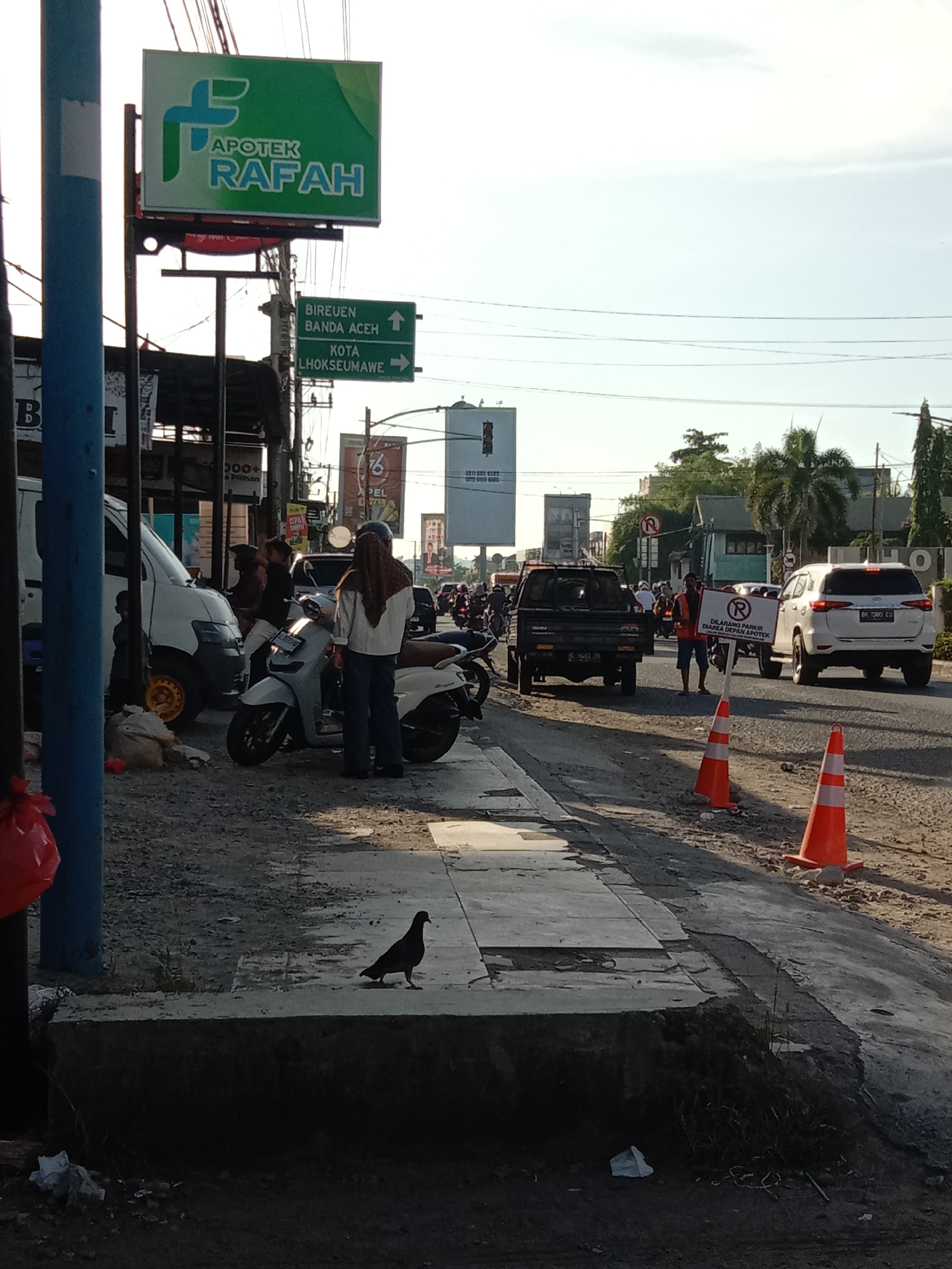 The image depicts a street scene in Lhokseumawe, Indonesia. A pigeon stands on the edge of a sidewalk in the foreground. Behind it, a person wearing a hijab stands next to a white scooter on a gravel shoulder. Further back, a white van, a black pickup truck, and a white SUV are visible among other vehicles on the road. Two orange traffic cones are placed near the road. A man in an orange vest holds a sign indicating "No Parking in the Pharmacy Area". Street signs for "Apotek Rafah" and directions to "Bireuen Banda Aceh" and "Kota Lhokseumawe" are visible. There are other commercial signs and billboards in the background, along with buildings and trees. The sky is bright, indicating it is daytime.
