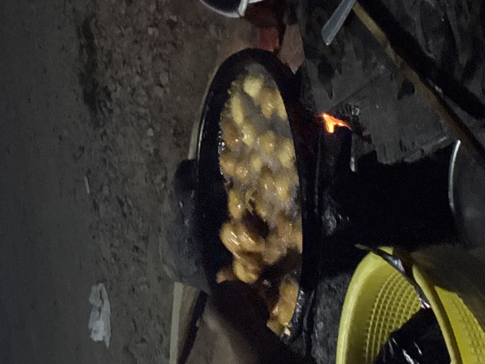 In Angwan Sayawa, Nigeria, a frying pan filled with dough balls is being heated over a small fire. The dough balls are golden brown and appear to be sizzling in hot oil. The pan is black and circular, and it is placed on a metal stand over a fire. The fire is small and orange, and it is producing some smoke. A yellow basket is visible to the left of the frying pan, and it contains some black plastic bags. The ground is covered in dirt and pebbles. The scene is dimly lit, suggesting it may be nighttime or early morning.