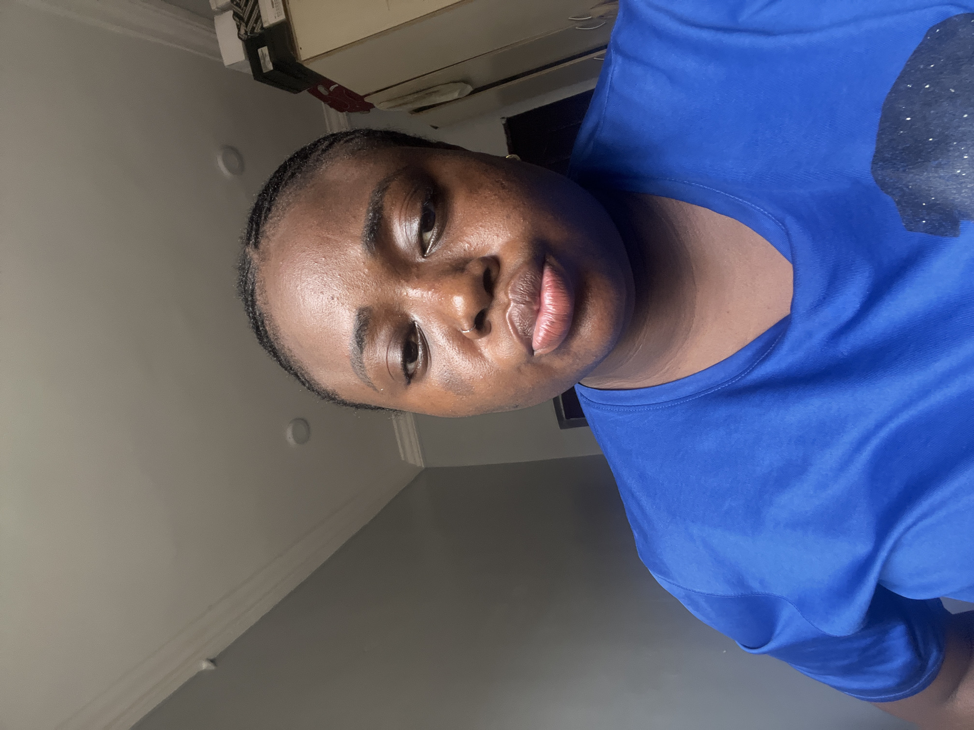 A close-up, slightly rotated portrait shows a woman with dark hair braided close to her head. Her skin tone is brown, and she is wearing a bright blue t-shirt. The background is comprised of a white ceiling with simple architectural molding, a light beige wall, and a gray wall. An out-of-focus object resembling a cabinet or shelf unit is visible in the upper portion of the frame. While details are limited, and further analysis would be needed for verification, it is implied from the request that the photo may have been taken in Kubwa, Nigeria. No specific landmarks or details are present to confirm this.