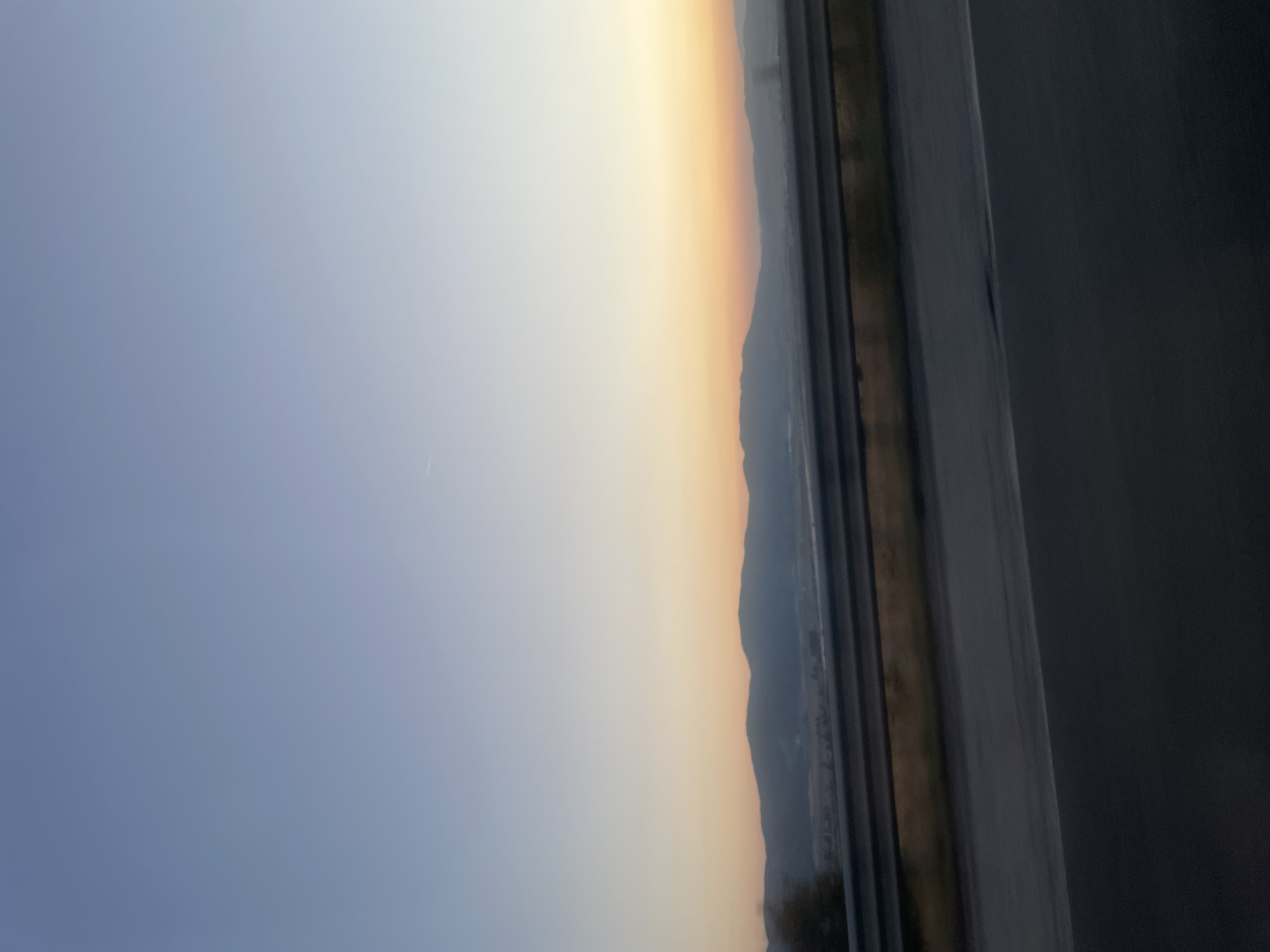 The image captures a serene natural landscape, likely taken from a moving vehicle, showcasing a broad expanse of the sky transitioning from a clear, light blue at the top to a warm, soft orange hue near the horizon. This gradient suggests either sunrise or sunset. In the middle ground, a range of dark, silhouetted mountains forms a distinct line against the colorful sky. Below the mountains, the landscape appears to be a valley or plain, with faint details of what could be fields or small settlements, partially obscured by atmospheric haze. A prominent horizontal element, a metallic guardrail, runs across the foreground, dividing the scene and suggesting the perspective is from a road. The lower part of the image shows the dark asphalt of the road, blurred by motion. The image does not contain any people, specific objects or actions, nor does it provide any explicit clues to the specific location within Ihtiman, Bulgaria, beyond the general description of a mountainous region with a road.