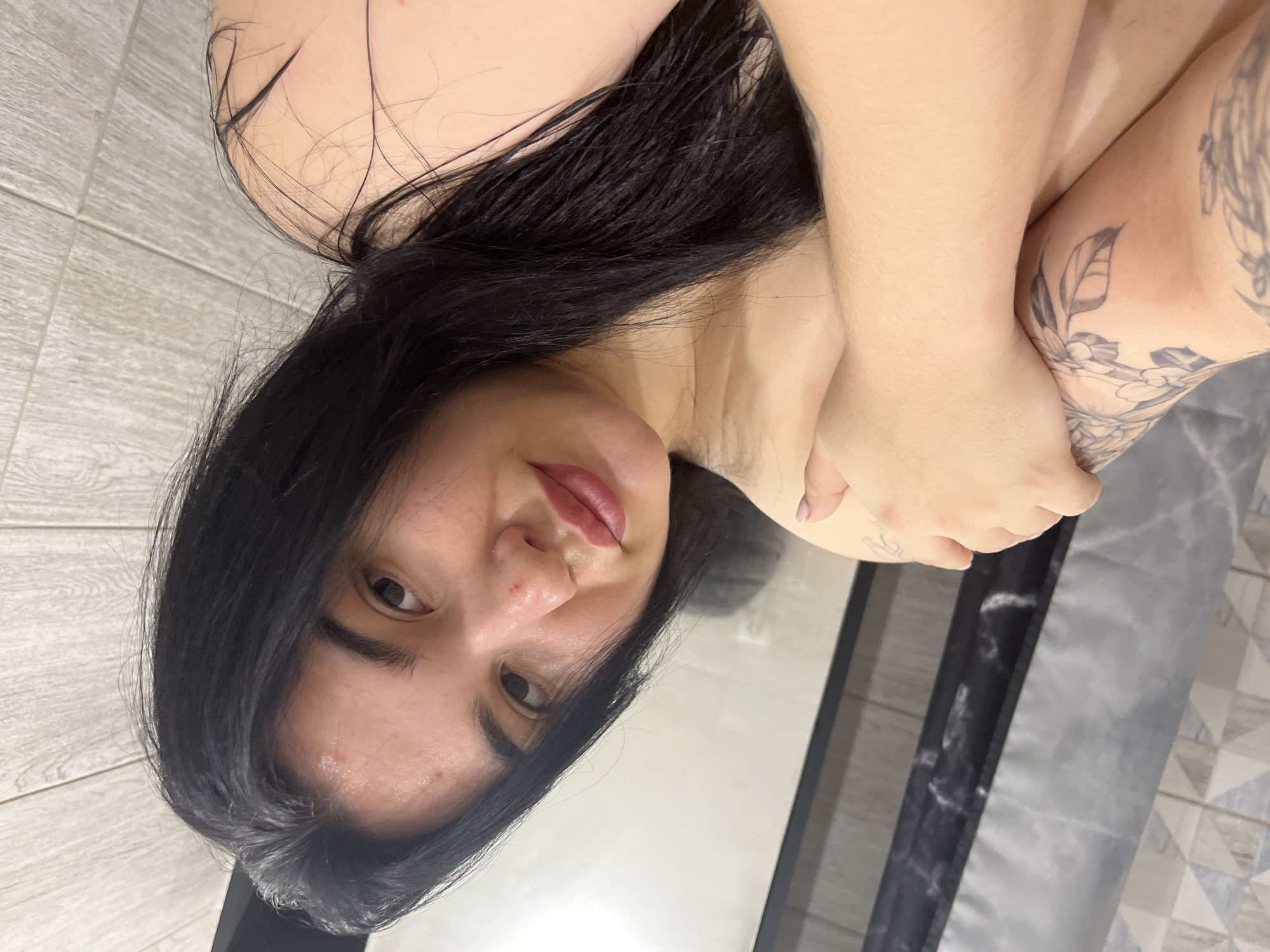 The image captures a close-up portrait of a person, seemingly taken from above, with the image itself rotated 90 degrees clockwise. The subject is a young-looking individual, likely female, with long, dark, slightly wavy hair that appears somewhat damp or tousled, spreading around their head and shoulders.
The person is looking directly at the camera with dark eyes and has a subtle, closed-mouth smile, showing reddish-pink lips that may be enhanced with lipstick or gloss. Their skin appears natural, with a few minor blemishes visible on the forehead and nose.
Their body is partially visible, showing light-toned skin on the shoulders and arms. One arm is bent, with the hand gently resting on the other arm, which is also bent at the elbow. A prominent tattoo of interconnected leaves and floral patterns in black and grey ink is visible on the upper part of their right arm, extending towards the shoulder. A small, indistinct dark mark or design is also visible on their left forearm.
The background on the left side of the image (which would be the bottom if rotated correctly) consists of a light-colored surface, possibly wood or ceramic tiles, featuring thin horizontal lines and a grid pattern. On the right side, there is a darker, patterned surface, possibly a bed or blanket, and a vertical dark strip that might be part of a furniture item or a mirror's edge, reflecting some light. The lighting appears soft and even, suggesting an indoor setting. There is no visible text in the image.