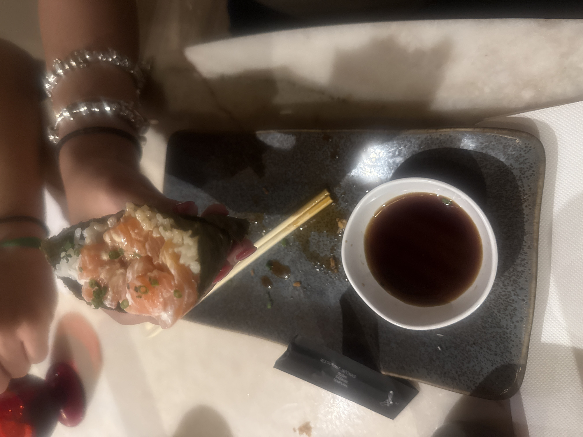 The image depicts a person holding a salmon temaki sushi roll, chopsticks, and a container of soy sauce on a grey rectangular dish. A dark chopstick wrapper with the words "RESTAURANT JAPONAIS SUSHI SASHIMI TEMPURA" is on the bottom of the plate. The background is blurry. The location is Loures, Portugal.