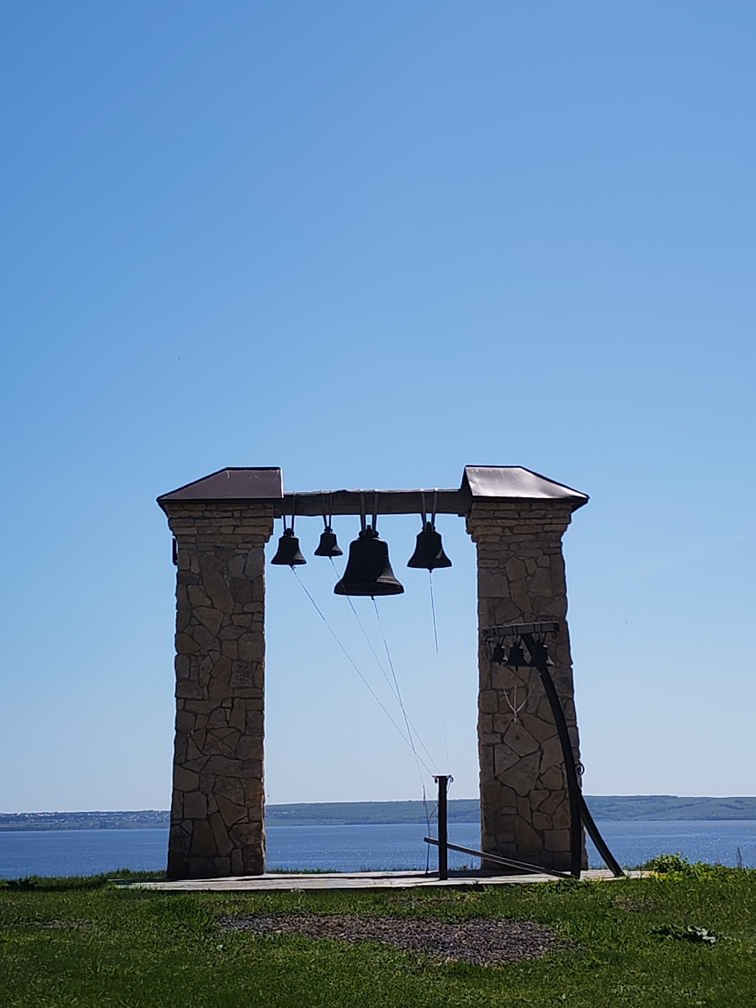 A medium shot captures a bell tower in Икское Устье, Russia, set against a clear blue sky and a distant body of water. The bell tower is constructed of two stone pillars, topped with dark roofing material, supporting a wooden beam from which several bells of varying sizes are suspended. Ropes for ringing the bells are visible. To the right of the tower, a metal structure with additional bells is leaning against one of the stone pillars. The scene is framed by green grass in the foreground, suggesting a park or open area. In the background, a body of water stretches across the horizon, with a shoreline visible on the opposite side.