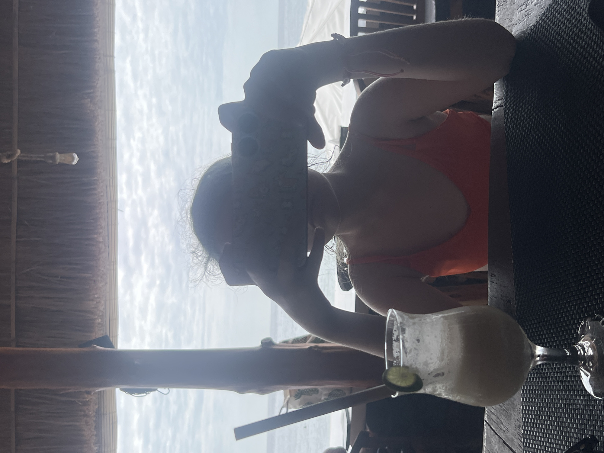 A young person, seen from the chest up, is seated at a table with a drink. The person holds a smartphone in front of their face, obscuring their features. They wear an orange top. The drink is in a stemmed glass with a lime wedge and a straw. In the background, a thatched roof structure is visible above a railing, beyond which the ocean and sky are seen. The setting appears to be a beachfront establishment.