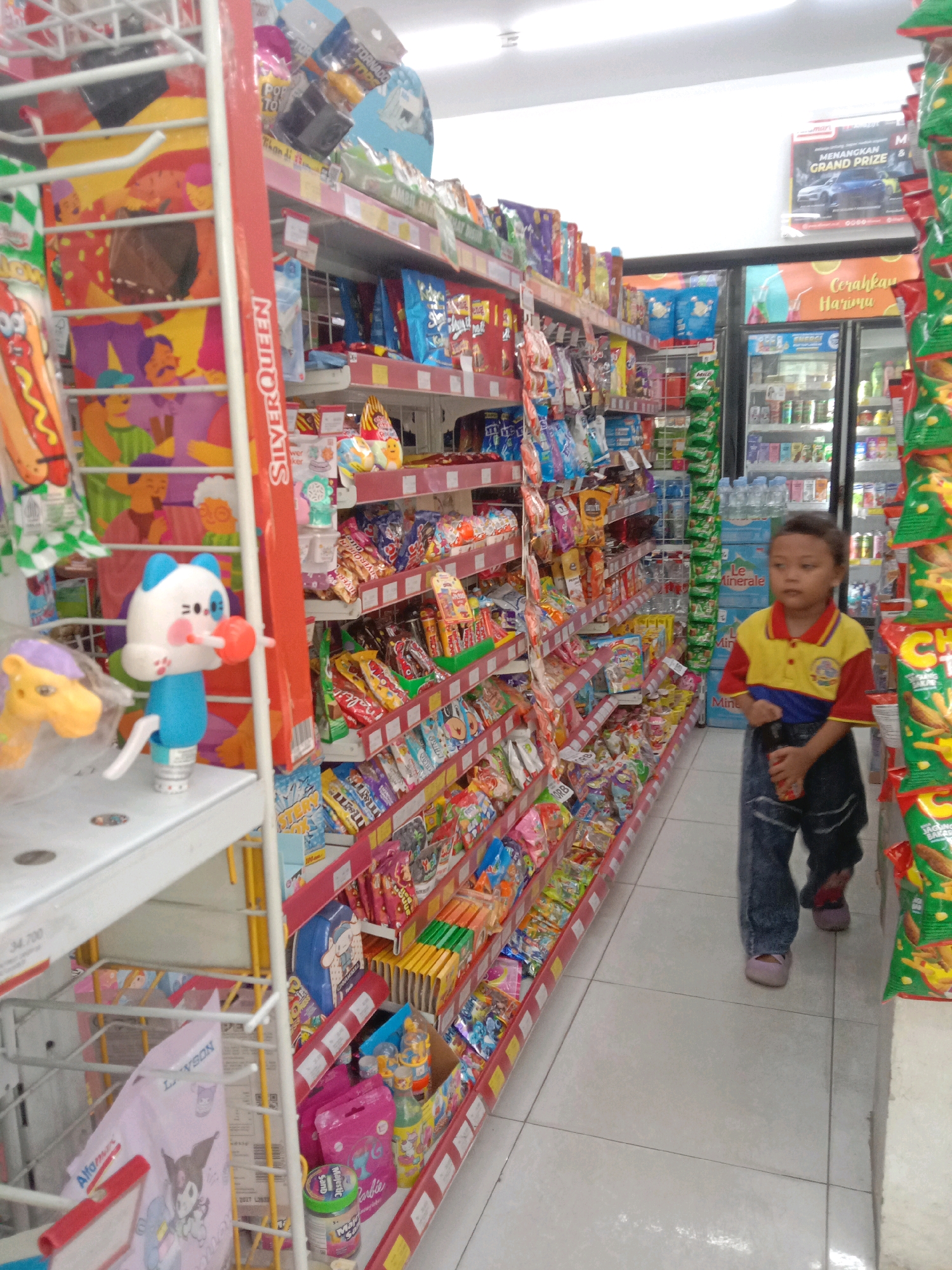 In a brightly lit convenience store, a young boy wearing a red and yellow polo shirt and blue jeans walks down an aisle stocked with various snacks and drinks. He is holding a small red object in his right hand. To his left, shelves are filled with colorful packaged goods, including bags of chips, candy, and what appear to be bubble bath products. In the foreground, a display rack holds novelty items, including a blue and white cat-shaped toy and a yellow horse toy. A refrigerator unit filled with beverages is visible in the background, along with a poster advertising a "GRAND PRIZE" and the text "Cerahkah Harimu" (Brighten Your Day). The floor is tiled with light-colored squares. The store appears to be a modern, well-organized retail establishment. This scene is consistent with a location in Karangraharja, Indonesia, which is known for its convenience stores and retail outlets catering to the local community.