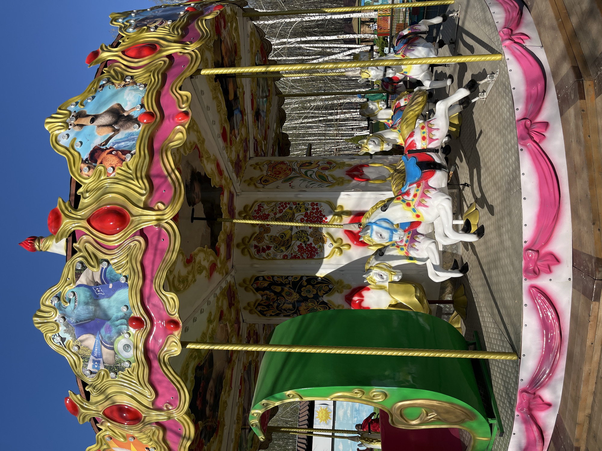 The image shows a colorful carousel in Chaikovsky, Russia. The carousel features ornate decorations with gold trim, pink accents, and framed images of cartoon characters. The carousel horses are white with painted saddles and bridles. In the background are birch trees. The sky is clear and blue.