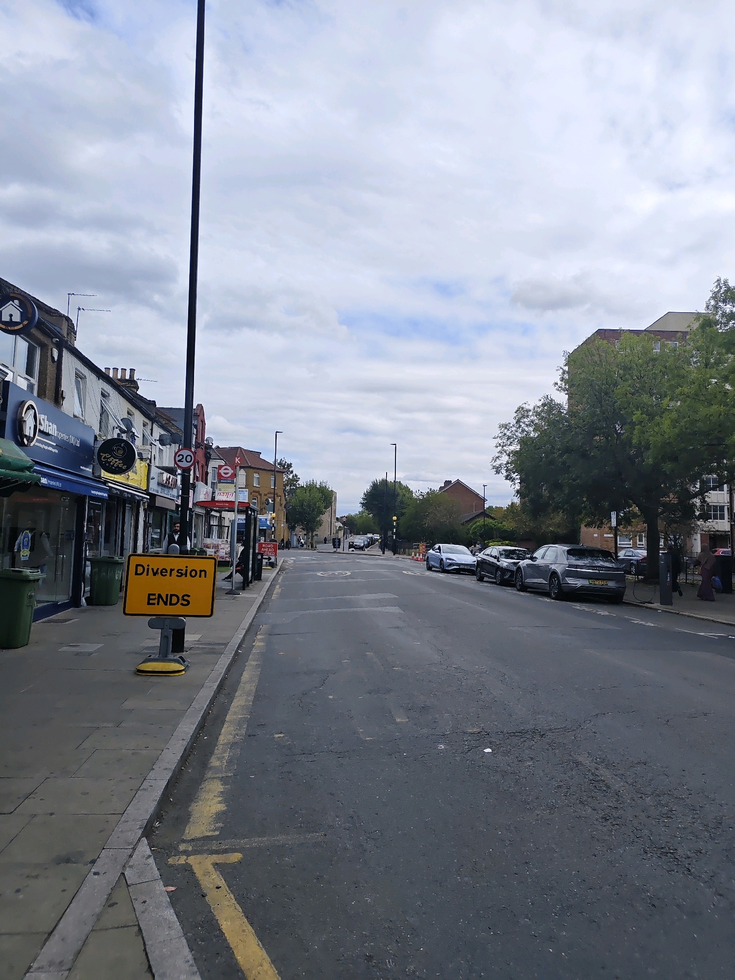 The image shows a street scene in London, United Kingdom. The foreground features a yellow diversion ends sign on a grey sidewalk. The sidewalk is bordered by a grey curb and a street with yellow lane markings and some potholes. To the left of the sidewalk are several shops, including one with a sign that says "Shan Properties". Green bins are visible next to the shops. On the street, cars are parked along the right side, and a few people are walking. Further down the street, a bus stop is visible, and traffic can be seen in the distance. The sky is cloudy.