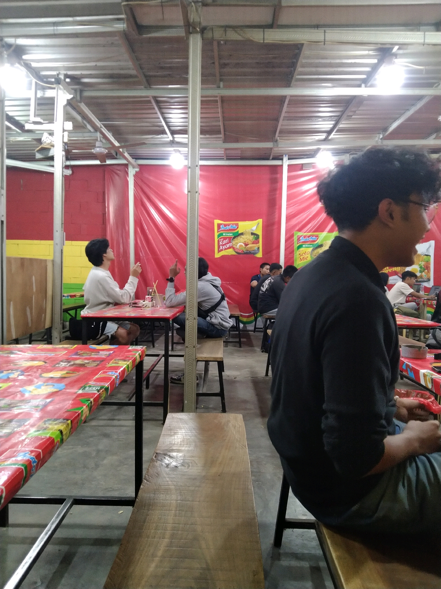 The image depicts an indoor food stall or restaurant setting in Malang, Indonesia. The establishment features a red and yellow color scheme on the walls and is covered by a corrugated metal roof supported by metal beams. Several tables with red tablecloths featuring various cartoon characters and food images are visible, along with wooden benches and chairs.
In the foreground, a person wearing a black long-sleeved shirt and grey pants is seated on a wooden bench, facing away from the camera. Their left arm rests on their lap, and their right arm is bent, with their hand near their face.
Further in the background, two young men are seated at a table. One, wearing a white long-sleeved shirt and dark shorts, is looking upwards and holding a cigarette. The other, wearing a grey hoodie, is gesturing with his right hand, which is raised.
Two posters, one for "Indomie Kari Ayam" (Indomie Chicken Curry) and another for "Indomie Soto Mie" (Indomie Beef Soto Noodle Soup), are prominently displayed on the red wall. Several other patrons are seated at tables in the background, some of whom are partially obscured. The floor is concrete. The overall atmosphere suggests a casual dining environment.