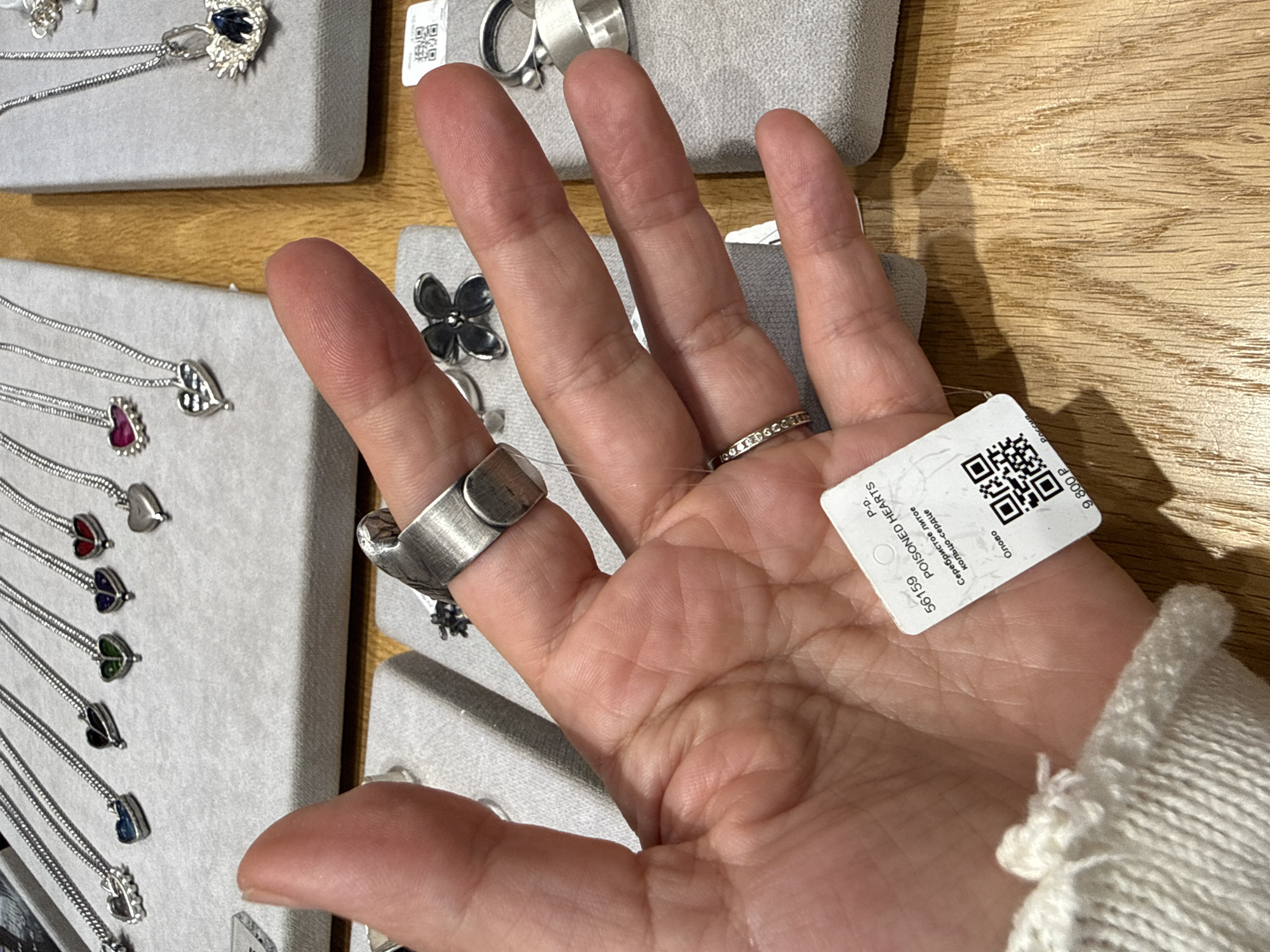 A close-up shot shows a person's left hand, which is wearing a silver ring with a textured surface and a smaller ring adorned with small stones on the ring finger. A tag is attached to the ring and reads "POISONED HEARTS". The background displays a collection of necklaces with heart-shaped pendants on display stands. The scene appears to be a retail environment, likely a jewelry store in Moscow, Russia, given the Cyrillic text on the tag. The wooden surface of a counter is visible at the bottom of the frame.