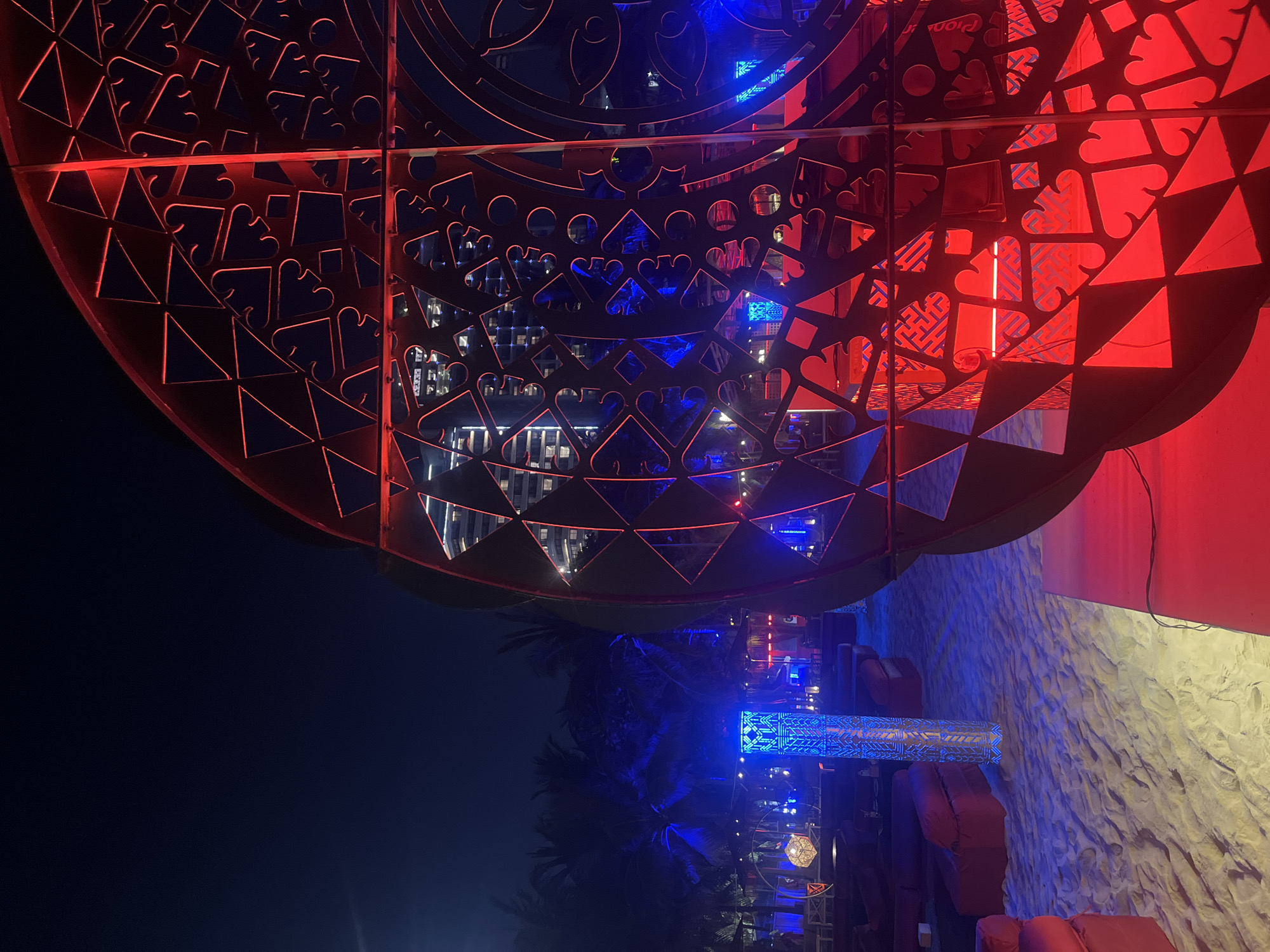 The image displays a vibrant outdoor or semi-outdoor lounge area at night, likely in Đà Nẵng, Vietnam, characterized by striking artificial red and blue lighting.
In the upper portion of the frame, a large, dark metal decorative screen or partition dominates, intricately laser-cut with a variety of patterns including hearts, triangles, and abstract geometric designs. This screen is dramatically illuminated from behind or within by a glowing red light, highlighting its detailed cutouts. Through these openings, the illuminated windows of distant buildings are visible, suggesting an urban or resort setting beyond.
The lower part of the image reveals the ground and other elements of the lounge. Tall palm trees are silhouetted against the dark night sky, partially lit by cool blue light. A prominent rectangular light pillar stands centrally with intricate blue geometric patterns. To the right, red cushioned seating, possibly sofas or benches, are arranged on a light-colored ground surface that appears to be sand. A textured, light-colored wall is also visible on the right, bathed in blue light. Another hexagonal light fixture can be seen near the bottom.
The overall atmosphere is modern, lively, and inviting, suggesting a sophisticated nighttime venue designed for relaxation or entertainment. The interplay of strong red and blue lights creates a dynamic and energetic ambiance. Faint text fragments, possibly "Pion" and "ra" from a brand name, can be seen on some reflective surfaces within the screen's cutouts.