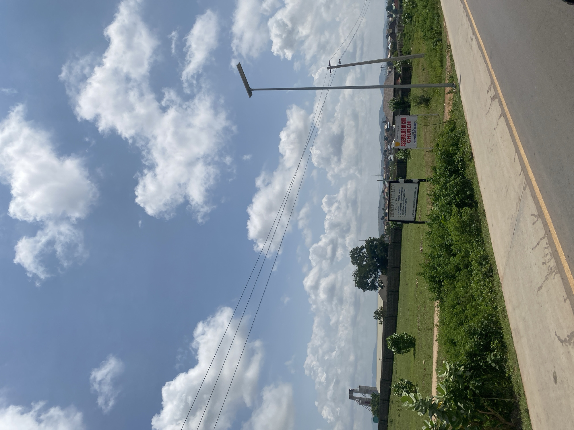 The image depicts a roadside view in Uke, Nigeria, under a bright, partly cloudy sky. In the foreground, a paved road with a yellow dividing line curves into the distance. To the left of the road is a concrete sidewalk and a grassy verge covered in lush green bushes. Further back, a flat, manicured lawn stretches towards a low brick wall. Beyond the wall, a cluster of buildings forms a village skyline, with hills visible in the background.
Several objects are notable in the scene. A tall, modern street light stands on the right side of the road. Further to the right, two utility poles with wires extend upwards. Two signs are visible in the grassy area. One sign, to the right, reads "ASSEMBLIES OF GOD CHURCH" with "UNITY (CITADEL OF GLORY)" below. Another sign to the left lists services from "CONCENTRY LTD." In the far left background, a tall, industrial-looking structure with a bridge-like design is prominent, possibly a construction site or an old factory. Scattered trees and smaller bushes dot the landscape. No people are visible in the image. The overall impression is one of a developing semi-urban or rural area.