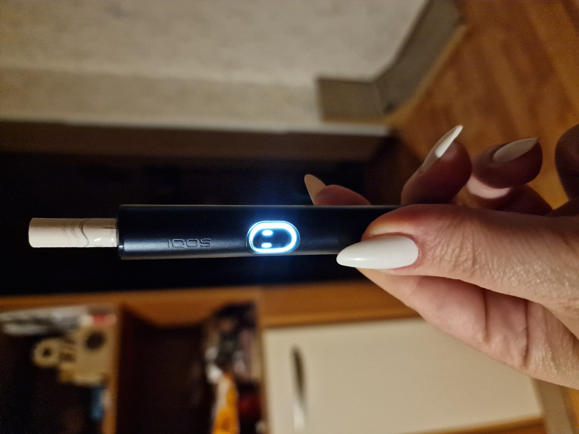 An extreme close-up shot shows a person's hand holding a black electronic smoking device with a light-up LED indicator. The LED displays a white smiley face icon. The device is inserted into a tobacco stick. The background is blurred, showing a shelf with items and a wall. The image appears to be taken indoors. There is no visible information to suggest the location is Pleven, Bulgaria.