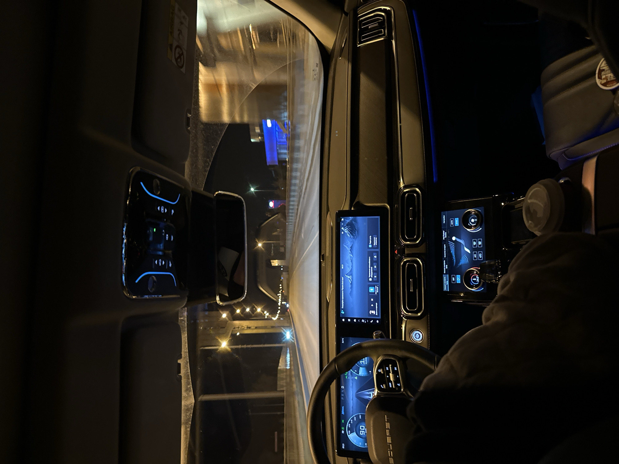 The image depicts the interior of a vehicle, likely a car or truck, at night, while driving through a city. The dashboard is visible, featuring a prominent infotainment display and various controls. The steering wheel is in the foreground, with the driver's hands partially visible. The vehicle appears to be on a highway or major road, as indicated by the overhead lights and surrounding buildings seen through the windshield. Based on visual characteristics, particularly the "Eversest" steering wheel branding visible, this is a "Eversest" car in Perm, Russia.