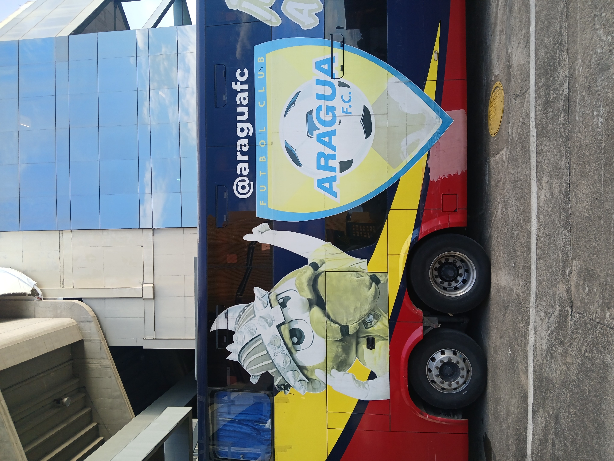 The image shows a bus with the logo of Aragua F.C., a Venezuelan football club. The bus is parked on a concrete surface, next to a modern building with glass windows. The bus is painted with the club's colors and features a cartoon mascot of the team. The location is likely Ciudad Guayana, Venezuela, given that Aragua F.C. is a Venezuelan club.